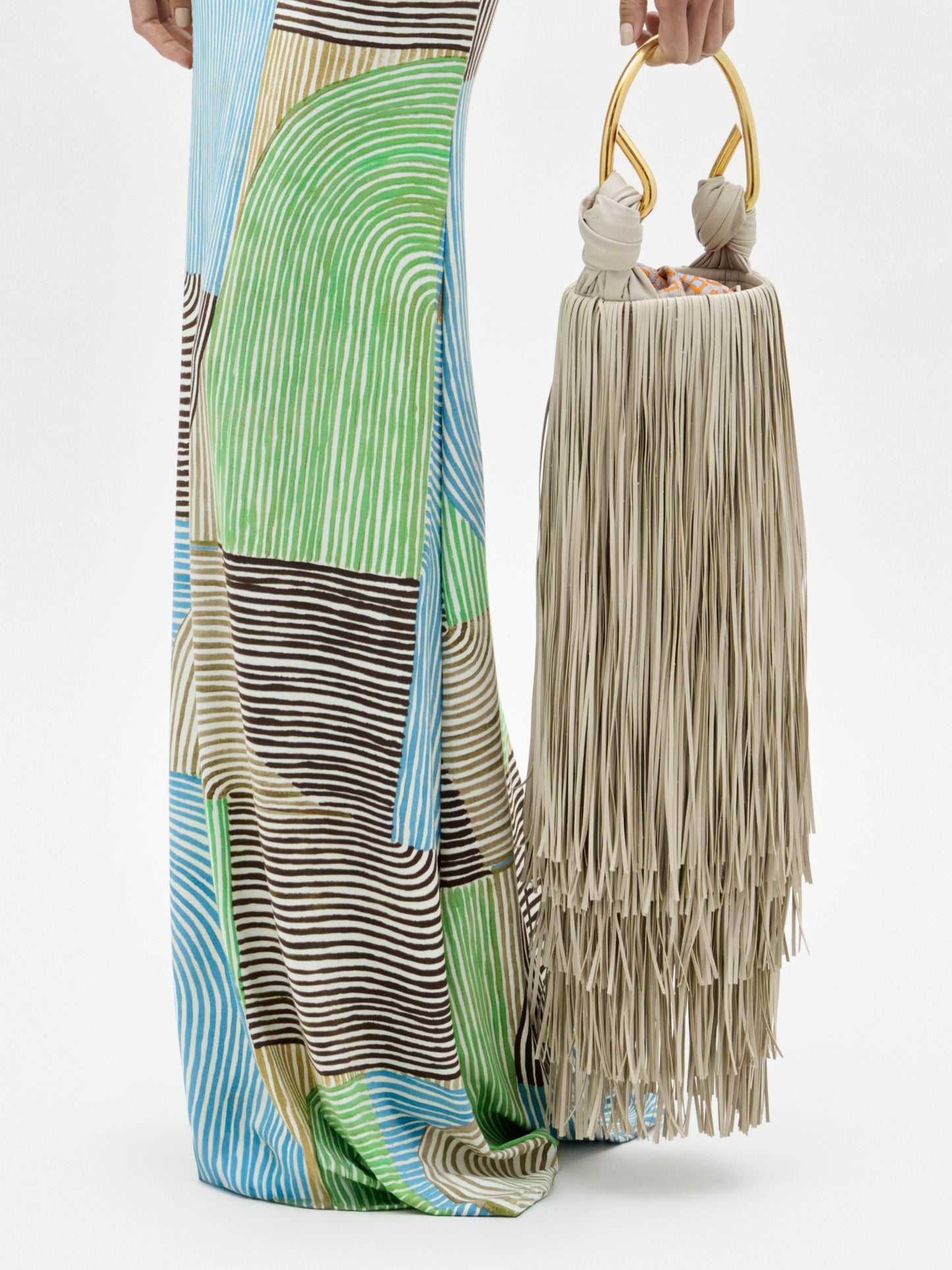 Una persona con un vestido a rayas de colores sostiene el Eugene Bag, un bolso beige con asas doradas y largos flecos en capas que casi tocan el suelo. Solo se ven la parte inferior del cuerpo y la mano.