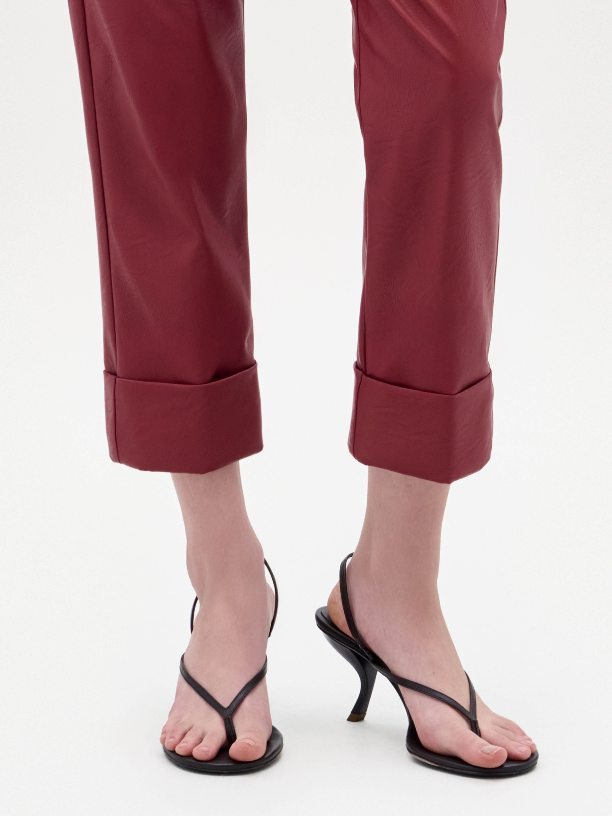 Una persona, de pie sobre un fondo blanco liso, lleva unos pantalones de cuero rojo enrollados y recortados y unos tacones Aurora negros.