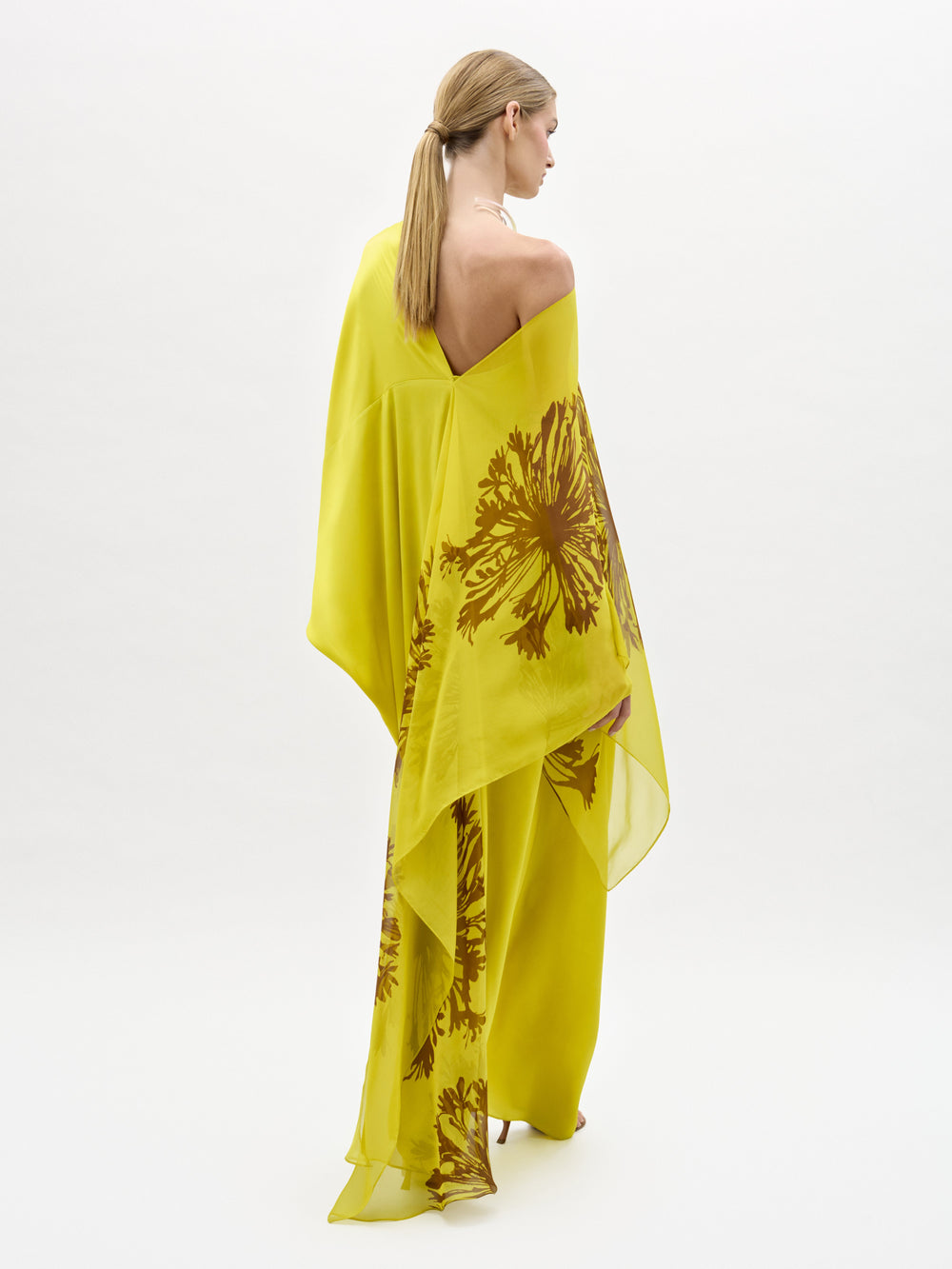 Una persona con el pelo rubio recogido en una coleta baja, de pie y de espaldas, lleva el Vestido de Jade, un maxivestido suelto de color amarillo semitransparente con motivos florales marrones, fabricado en Colombia. El fondo es blanco liso.