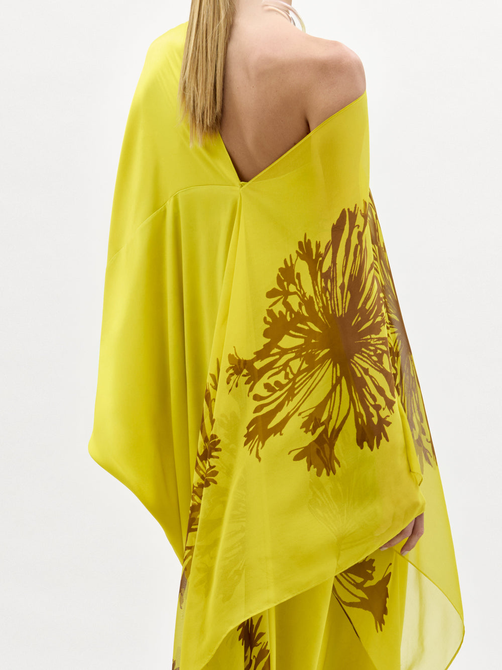 Una persona de pelo largo y rubio lleva el Vestido de Jade, un maxi amarillo brillante de un solo hombro con motivos florales marrones. La imagen muestra la espalda y parte del brazo izquierdo sobre un fondo blanco liso.