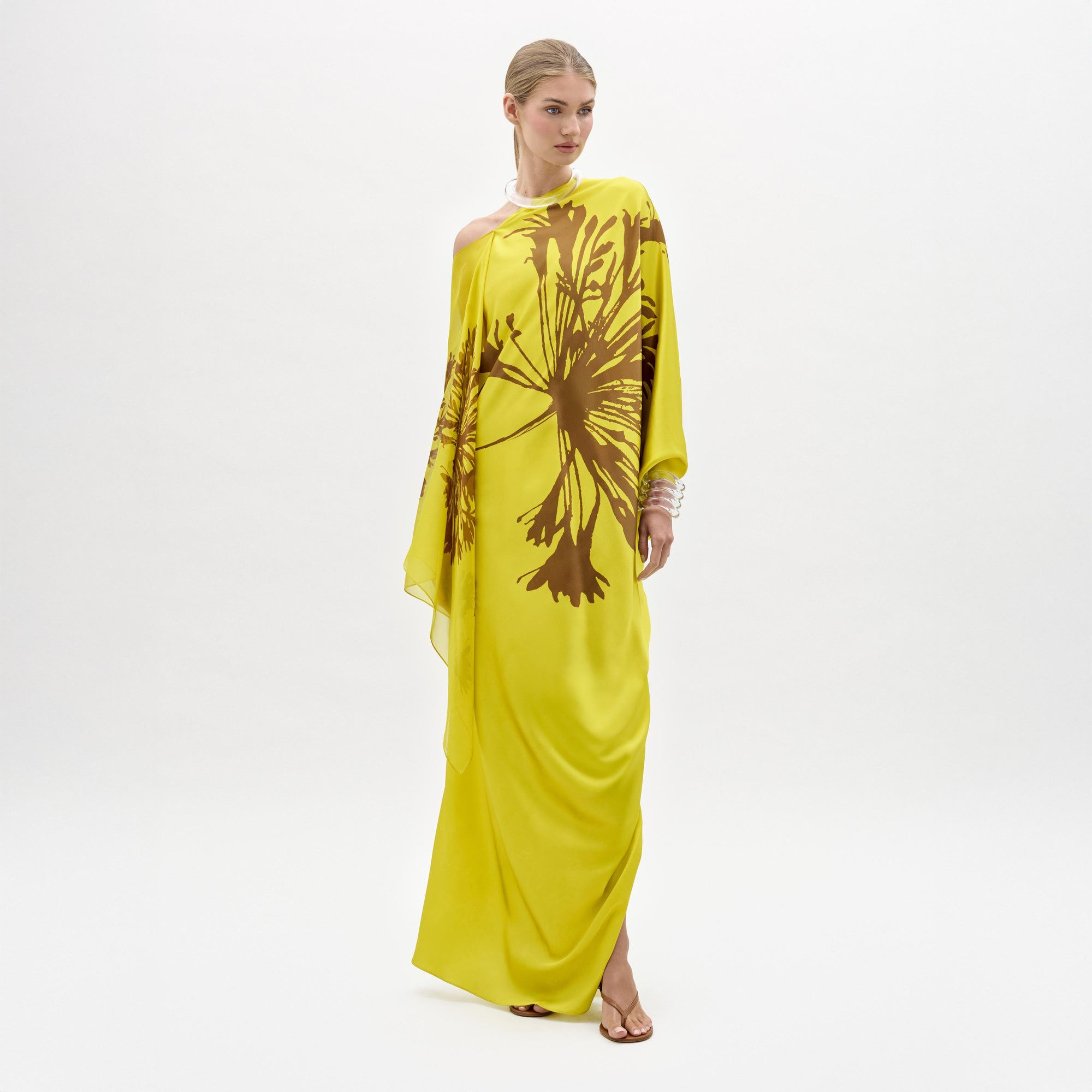 Una persona sobre fondo blanco luce el vestido Jade -un maxi amarillo brillante de un solo hombro con llamativos motivos florales marrones en tejido italiano- combinado con sandalias planas. Fabricado en Colombia.