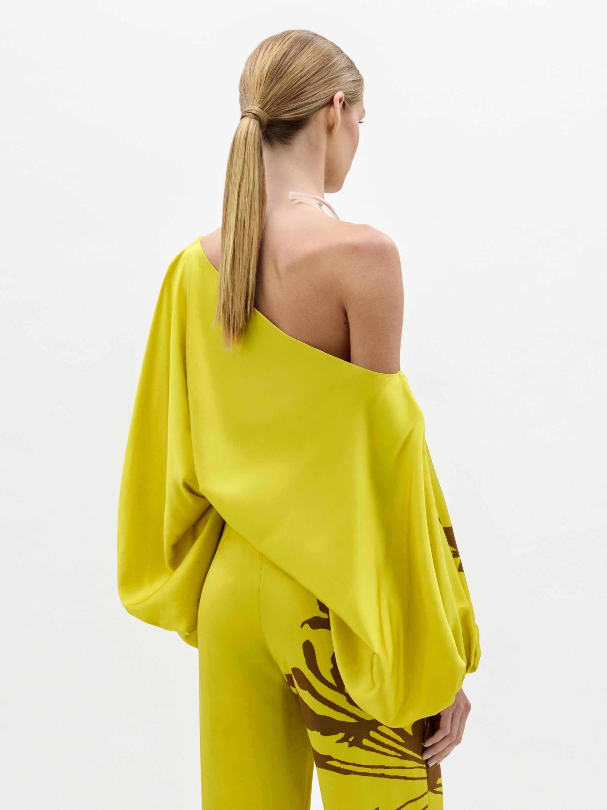 Una mujer con el pelo rubio liso recogido en una coleta baja lleva la blusa Bellagio, un top amarillo brillante sin hombros con mangas abullonadas, combinado con unos pantalones marrones de estampado abstracto a juego. Está de espaldas sobre un fondo liso.
