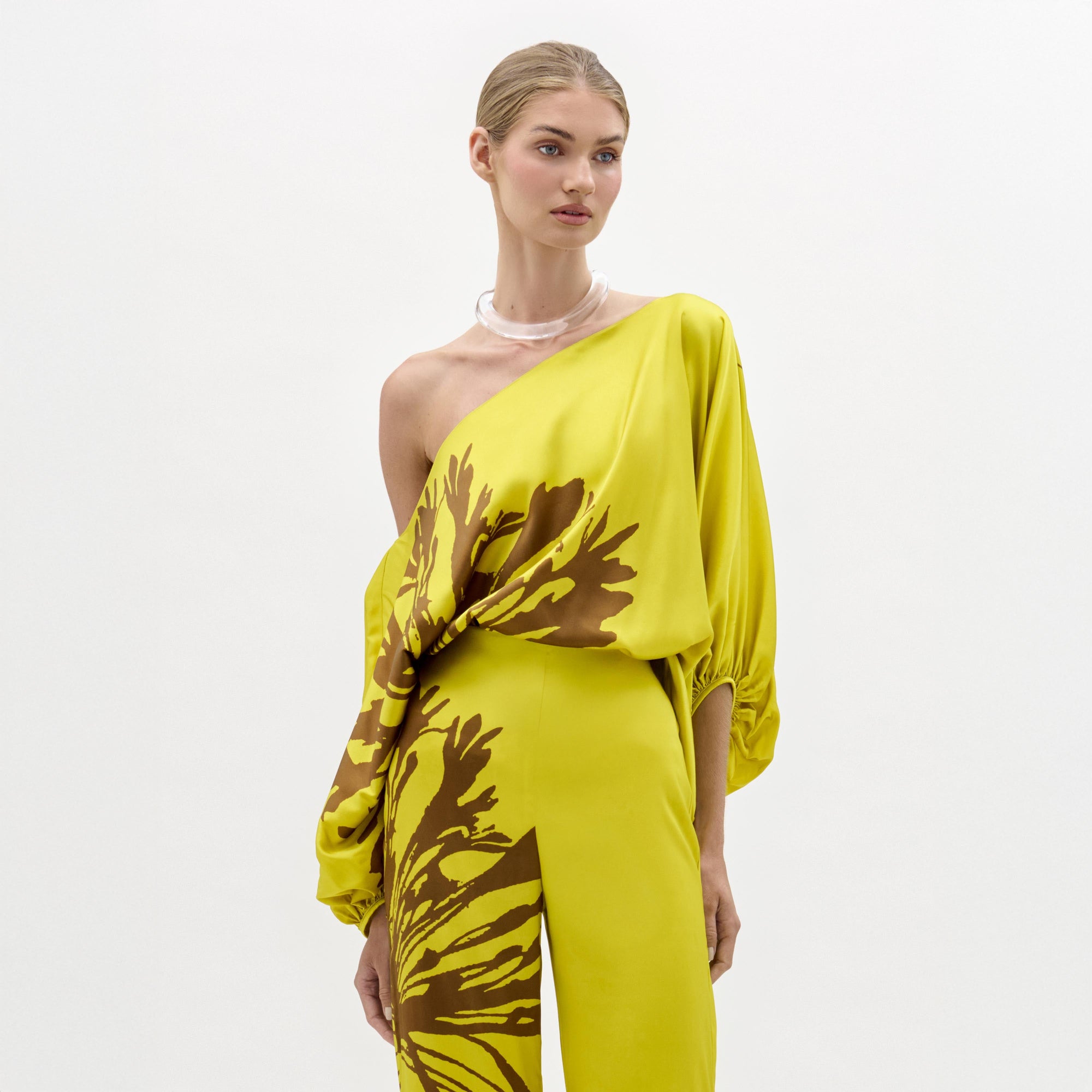 Una mujer sobre un fondo liso luce la blusa Bellagio, un top amarillo brillante de un solo hombro con motivos abstractos marrones en tela italiana. Lleva el pelo claro recogido hacia atrás y un collar transparente y grueso como accesorio.