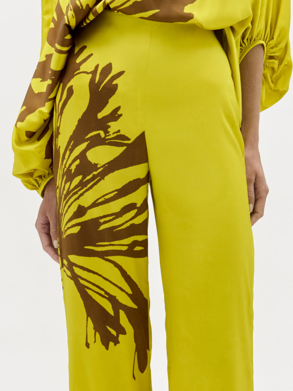 Una persona lleva el pantalón Andie Pant, de color amarillo brillante y cintura alta, con un llamativo estampado floral abstracto marrón en una pierna, combinado con un top amarillo y marrón a juego. Sus manos descansan a los lados sobre un fondo blanco liso.
