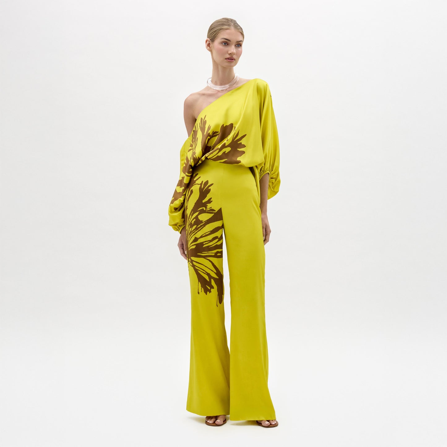 Sobre un fondo blanco, una persona luce el Andie Pant -un mono amarillo brillante de un solo hombro confeccionado en tela italiana con flores abstractas marrones y perneras anchas- con sandalias de tacón, el pelo recogido y un collar.