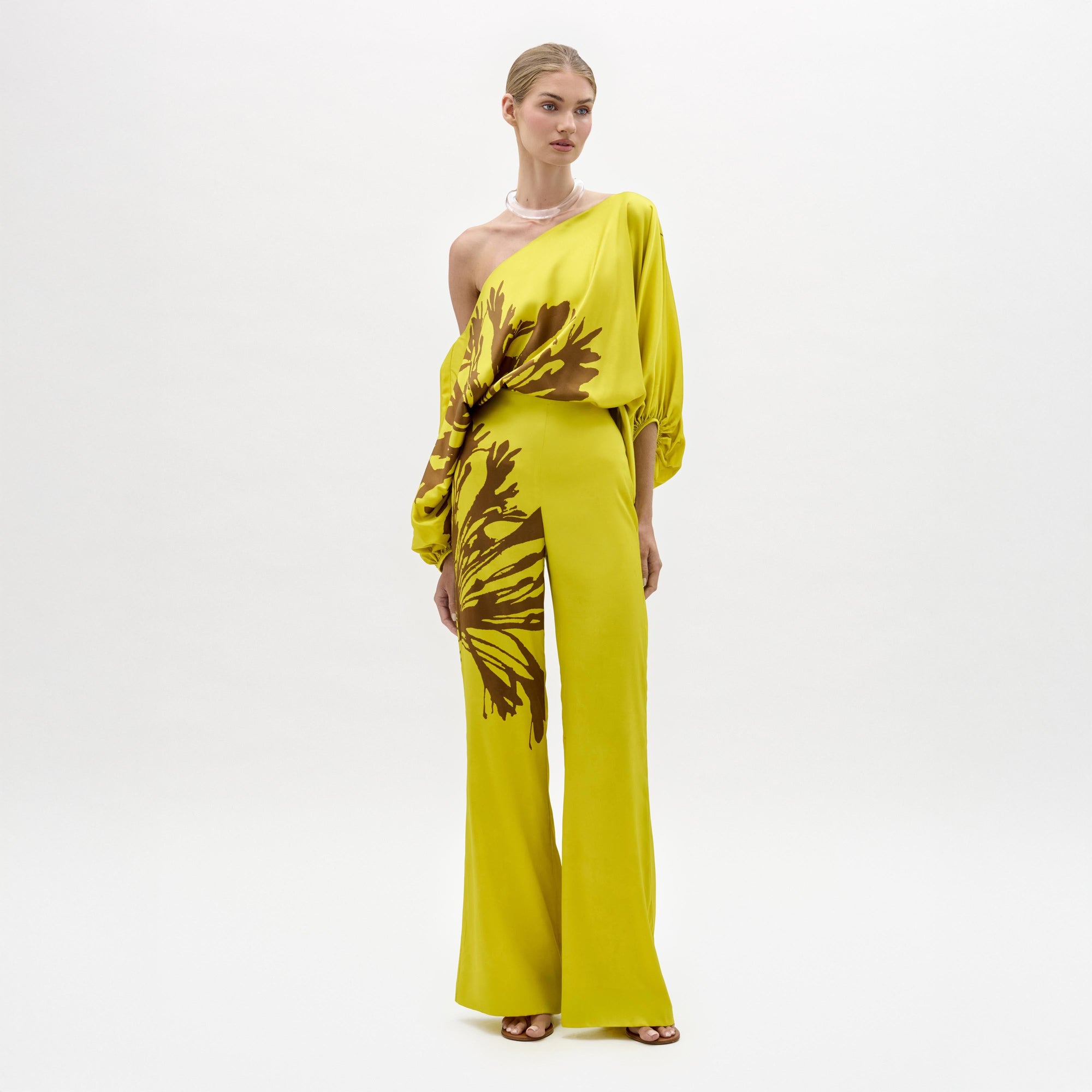Sobre un fondo blanco, una persona luce el Andie Pant -un mono amarillo brillante de un solo hombro confeccionado en tela italiana con flores abstractas marrones y perneras anchas- con sandalias de tacón, el pelo recogido y un collar.