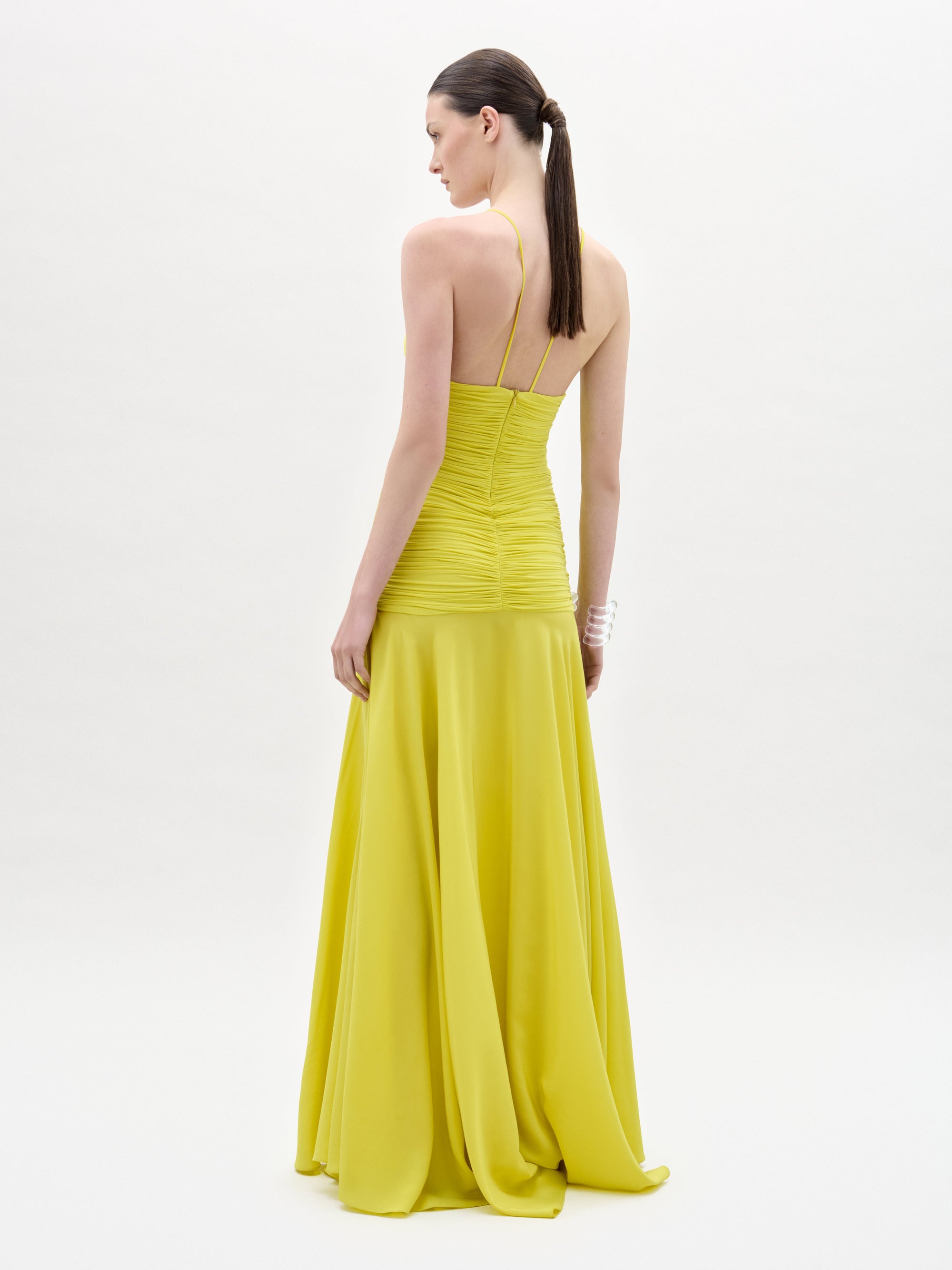 Una mujer mira hacia otro lado y lleva el vestido Sana de color amarillo brillante, largo hasta el suelo, con tirantes finos y corpiño fruncido. Lleva el pelo liso recogido en una coleta baja y un brazalete blanco en la muñeca izquierda.