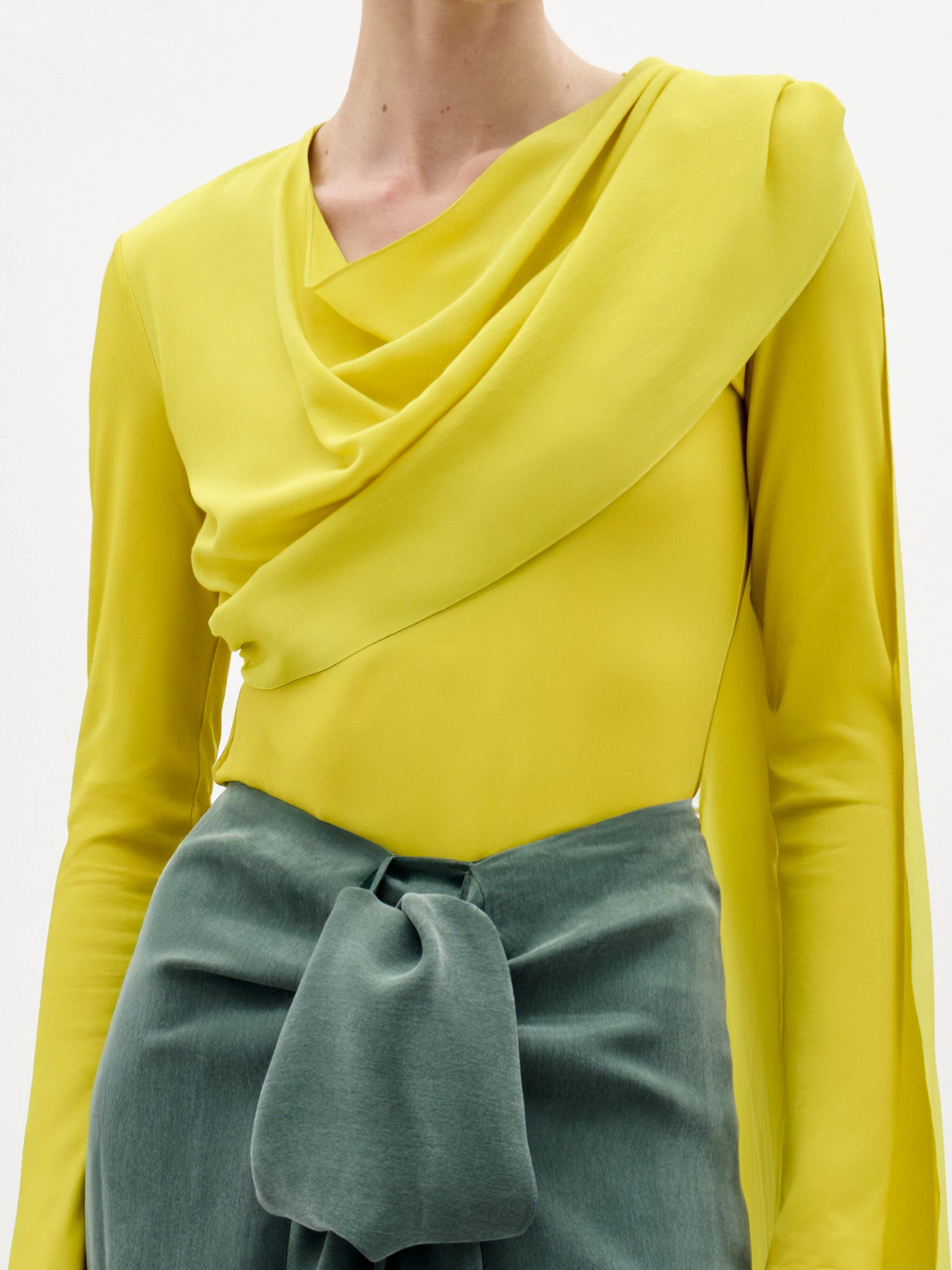 Una persona lleva la blusa Josic, un top asimétrico amarillo de manga larga y drapeado, con unos pantalones verde salvia de talle alto con un gran lazo delantero; la imagen muestra sólo la parte superior de su cuerpo.