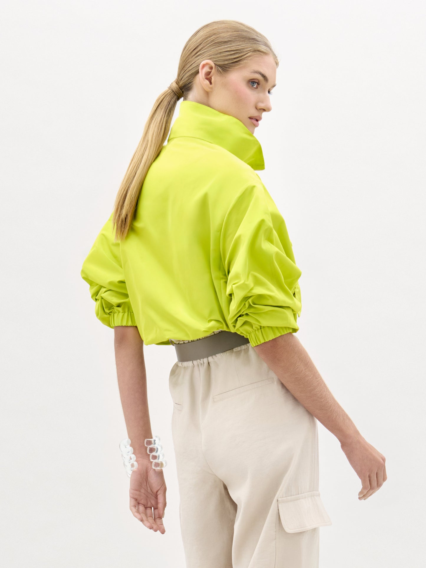 Una persona con el pelo largo y rubio recogido en una coleta baja lleva la chaqueta Donata -de color verde brillante, de cuello alto y talla grande, con mangas fruncidas- combinada con unos pantalones cargo de cintura alta de color beige claro. Está girada hacia un lado y lleva una pulsera blanca gruesa.