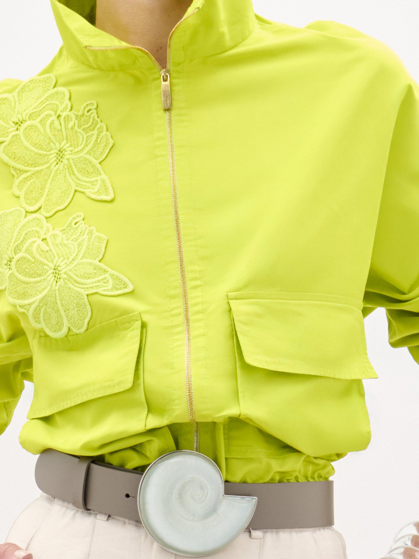 Una persona viste la Chaqueta Donata, una chaqueta corta de gran tamaño de color amarillo verdoso brillante con grandes bolsillos delanteros y bordados florales en un lado, que se combina con pantalones claros, un cinturón gris y un accesorio verde claro en forma de nautilus en el cinturón.