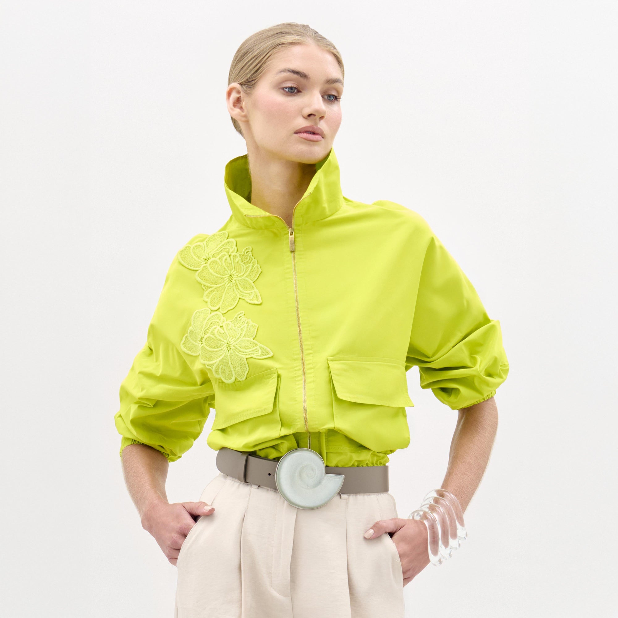 Sobre un fondo blanco liso, una persona viste la chaqueta Donata de color amarillo verdoso brillante con bordados florales, pantalones de cintura alta de color beige, una gran hebilla de cinturón circular y un brazalete transparente.