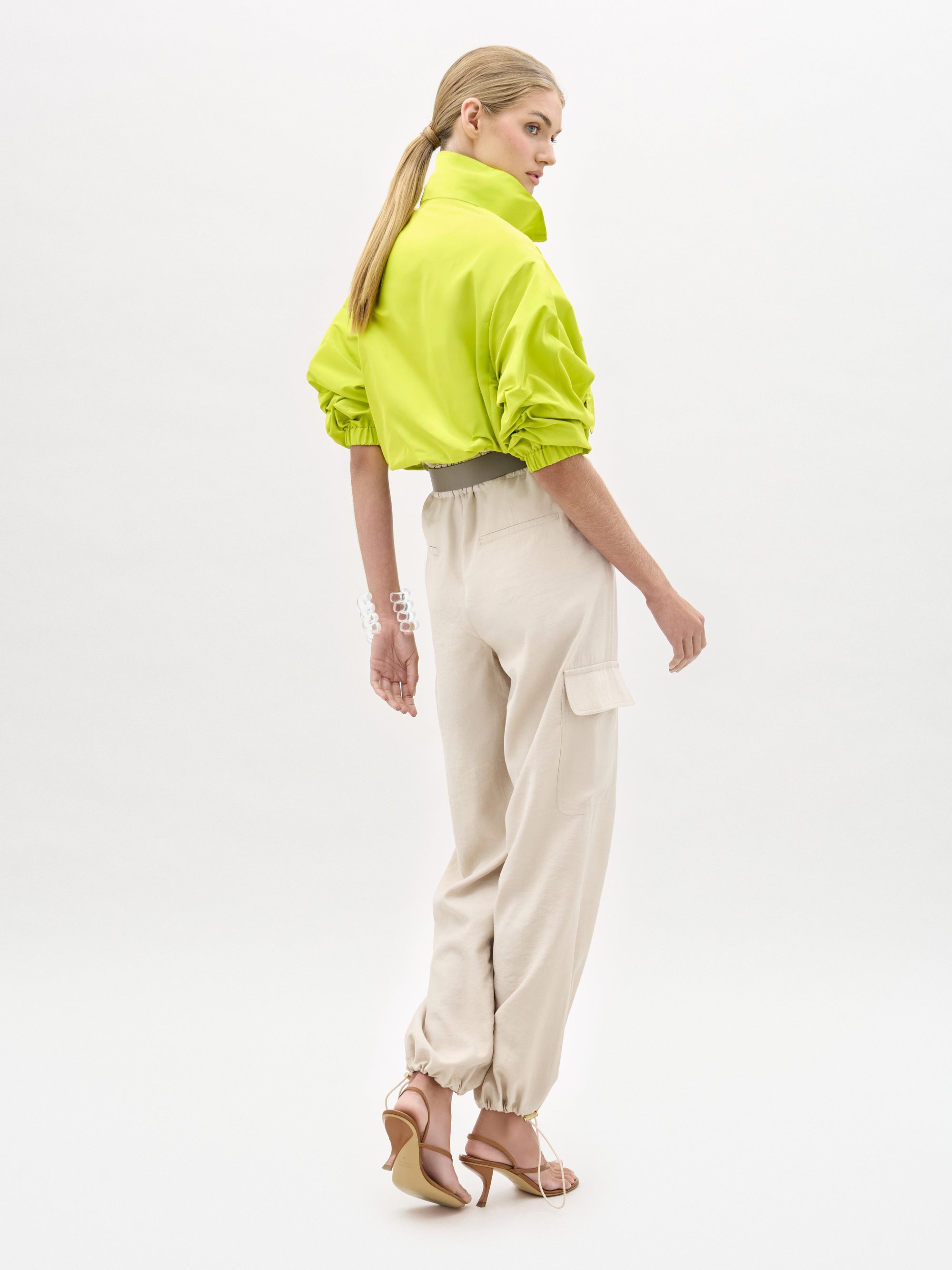 Una persona con el pelo largo y rubio recogido en una coleta baja lleva una chaqueta recortada de color amarillo brillante, el pantalón Darnell de estilo cargo beige de tiro alto y sandalias de tacón rosas, de pie ante un fondo blanco y mirando por encima del hombro.