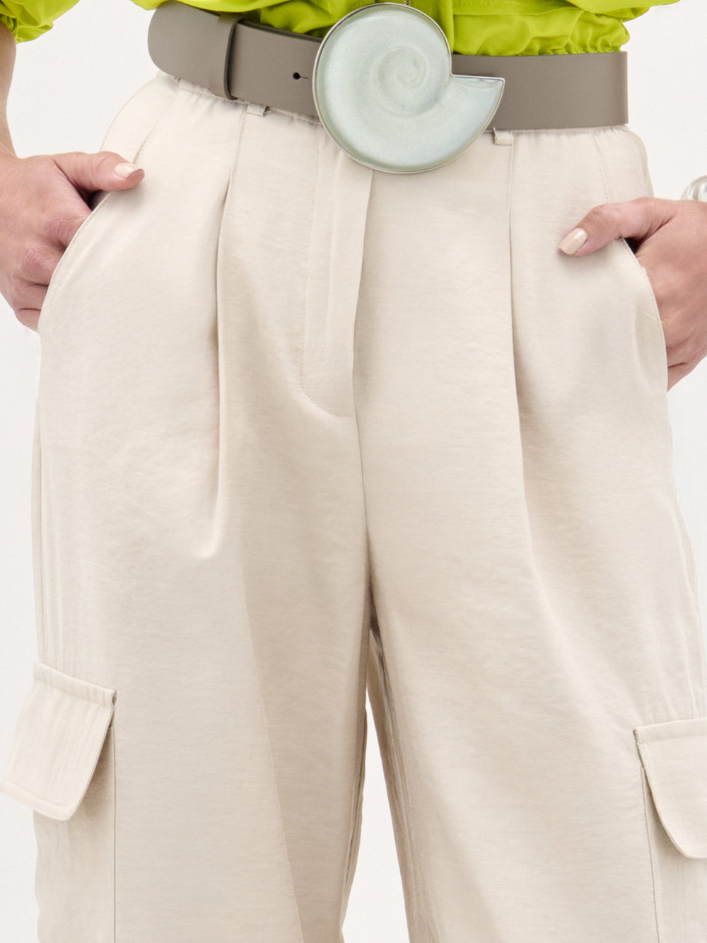 Una persona lleva el Darnell Pant-beige cargo de talle alto con bolsillos laterales y un cinturón ancho gris con una gran hebilla en espiral. Las manos en los bolsillos, combinado con una camisa verde lima. Hecho en Colombia.