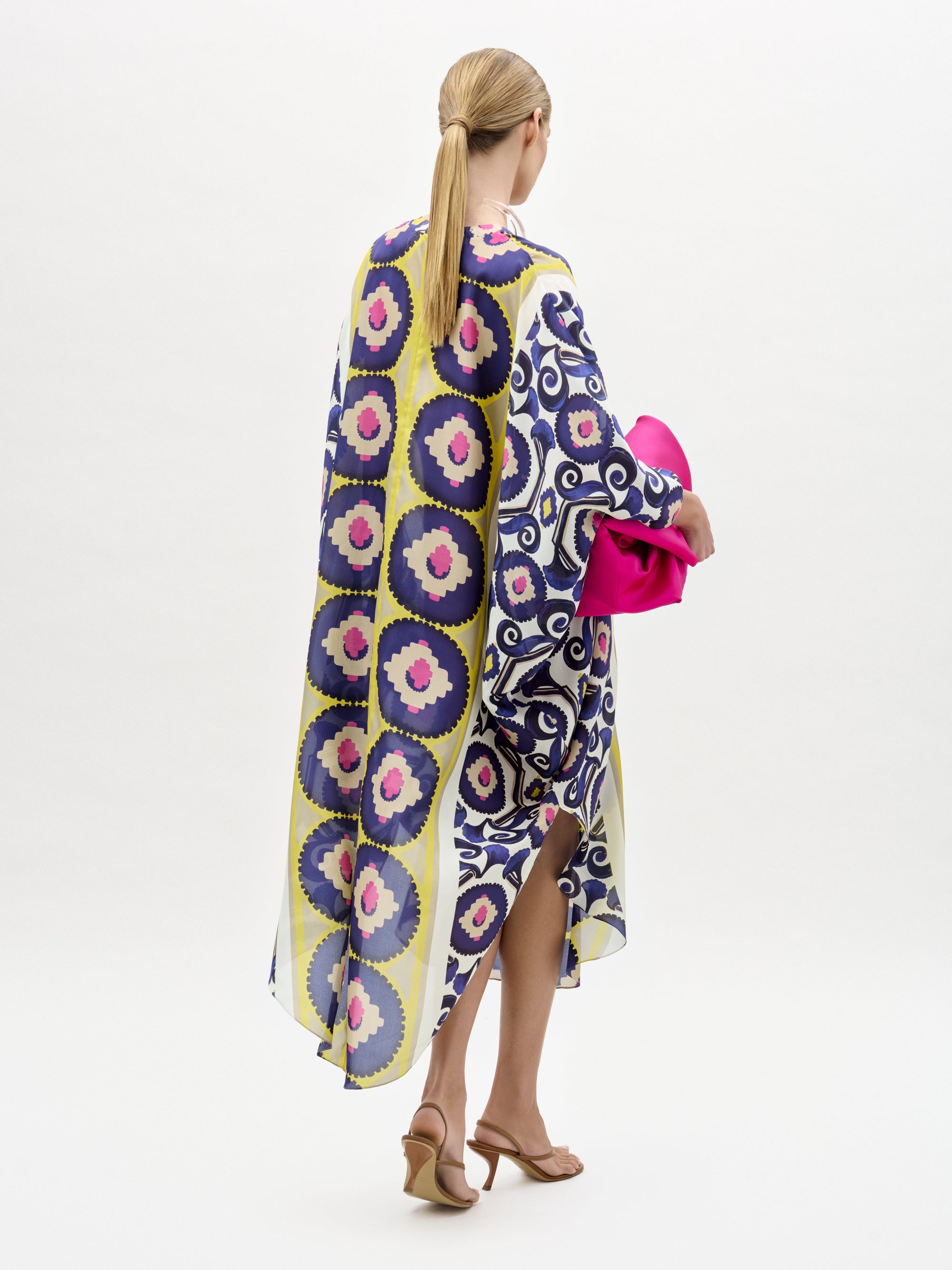 Una mujer con el pelo rubio recogido en una coleta baja lleva la túnica Ilse, un vestido largo suelto con motivos geométricos y florales. Lleva un bolso de mano rosa brillante y sandalias de tacón color canela sobre un fondo blanco liso.