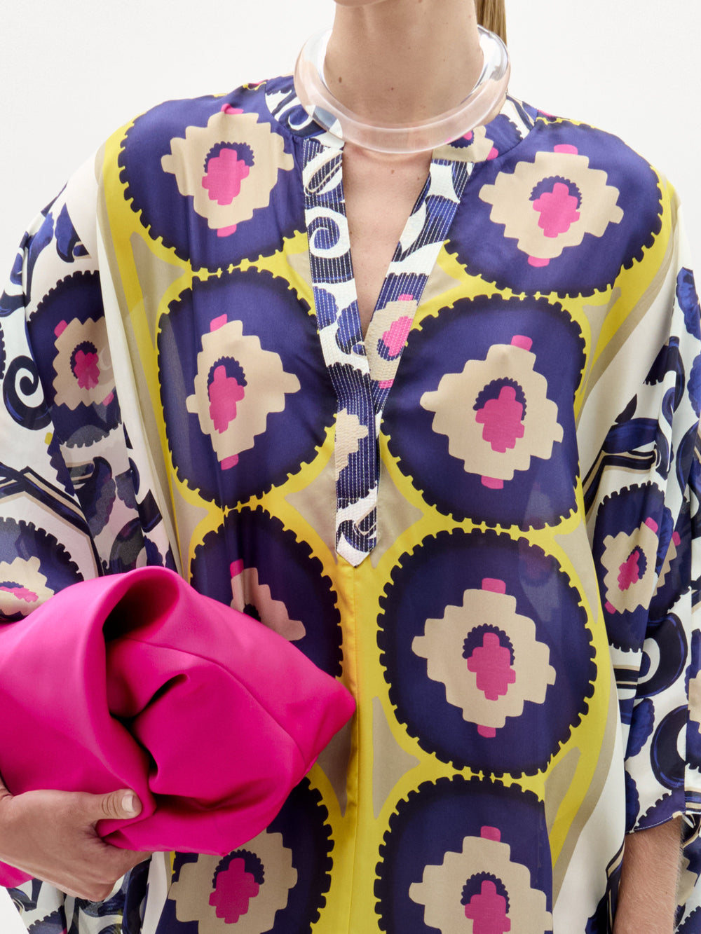 Una persona lleva la túnica Ilse, un maxivestido hecho en Colombia con diseños circulares en azul, amarillo y rosa, y sostiene un clutch rosa brillante. El escote abierto se combina con un collar transparente.