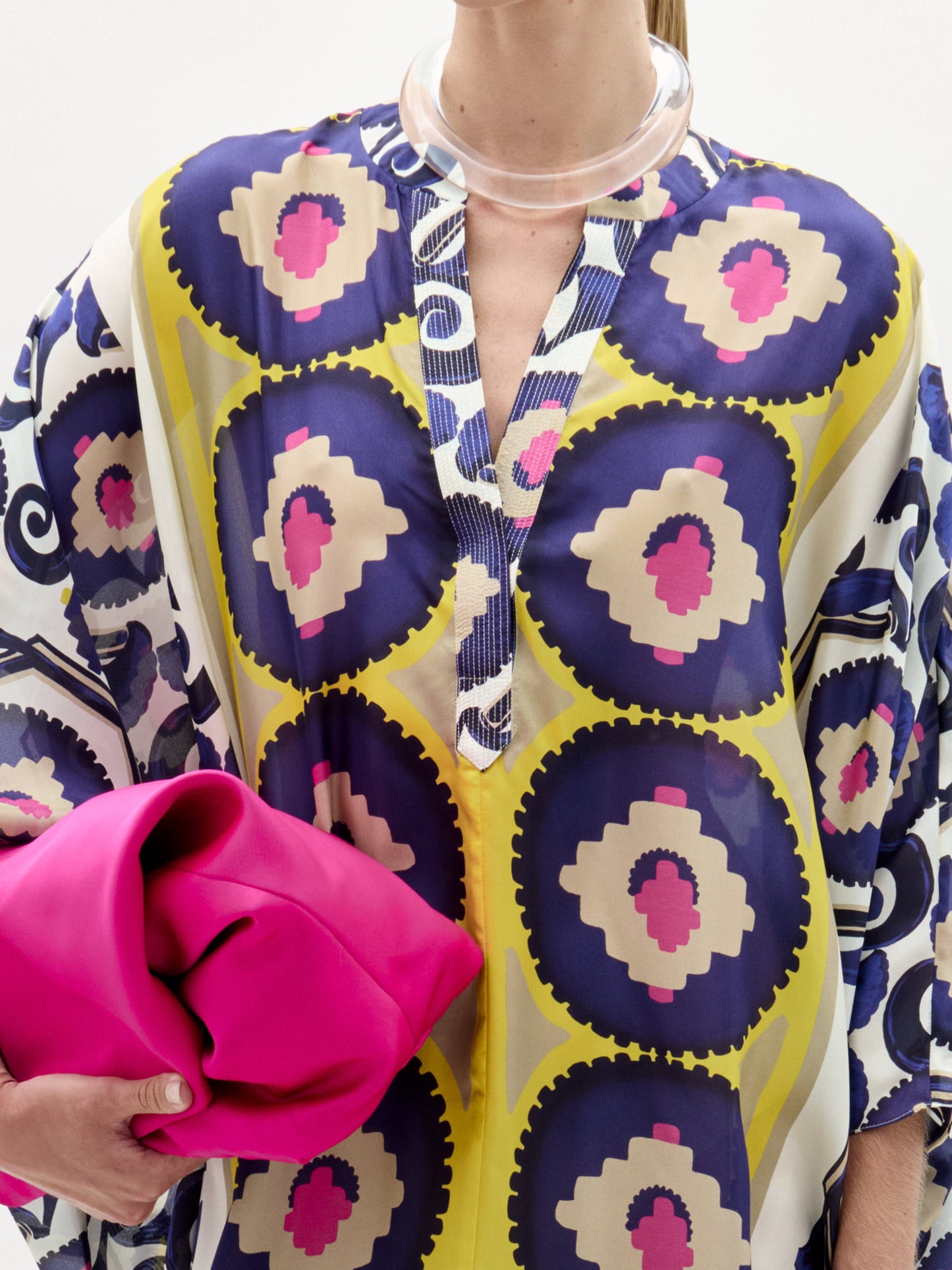 Una persona lleva la túnica Ilse, un maxivestido hecho en Colombia con diseños circulares en azul, amarillo y rosa, y sostiene un clutch rosa brillante. El escote abierto se combina con un collar transparente.