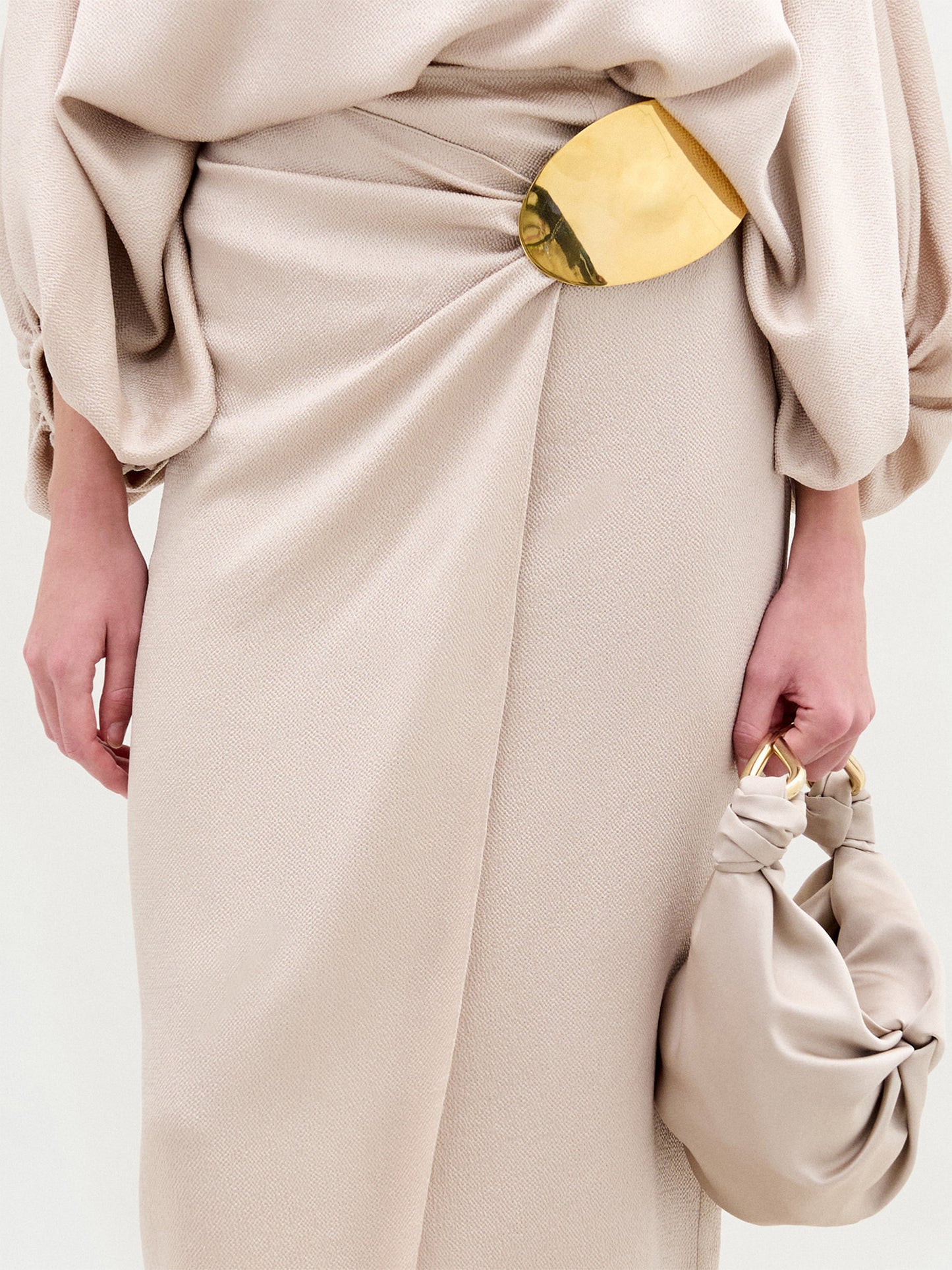 Una persona lleva la Falda Adrianne, una falda martillada de satén beige con tejido drapeado y un gran adorno ovalado dorado en la cintura, que sostiene un bolso anudado beige a juego. Sólo se ven el torso y las manos. Hecho en Colombia.