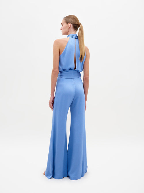 A person wearing the Siv Jumpsuit Periwinkle, featuring a sleeveless, light blue, halterneck design with wide-leg pants and a belted waist, stands against a plain white background. Pre-order now to ensure it ships by February 15th, 2025.