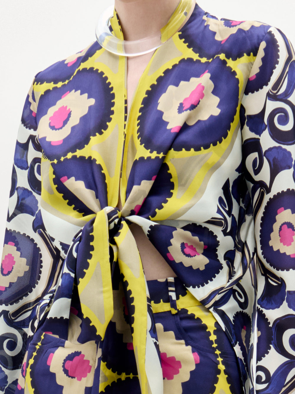 Una persona lleva la Blusa Miel, un vibrante top de manga larga confeccionado en 100% seda con motivos circulares en azul, amarillo, rosa y beige. Lleva un nudo delantero y cuello de pico, y se combina con unos pantalones de cintura alta a juego. No se ve la cara.