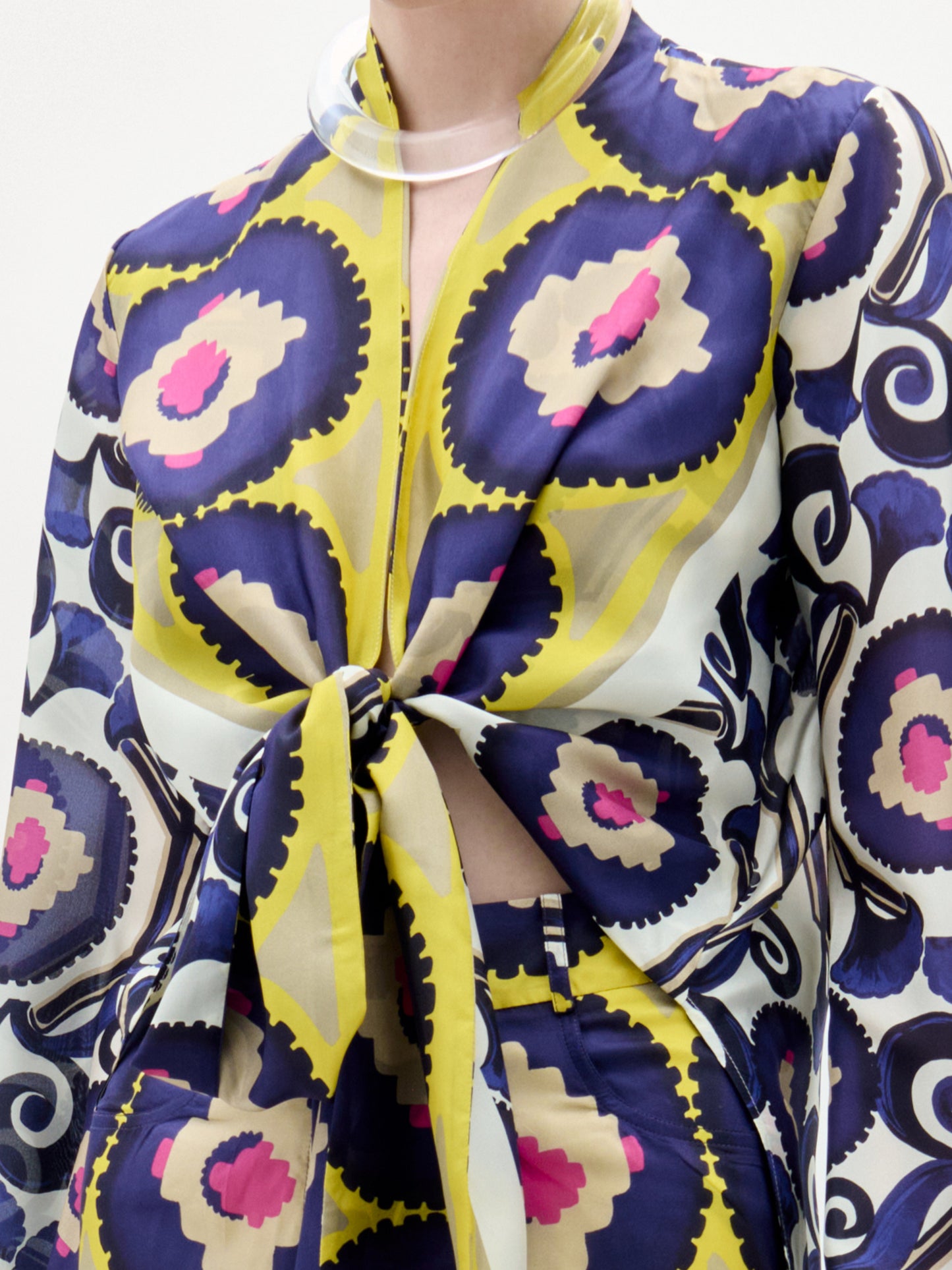 Una persona lleva la Blusa Miel, un vibrante top de manga larga confeccionado en 100% seda con motivos circulares en azul, amarillo, rosa y beige. Lleva un nudo delantero y cuello de pico, y se combina con unos pantalones de cintura alta a juego. No se ve la cara.