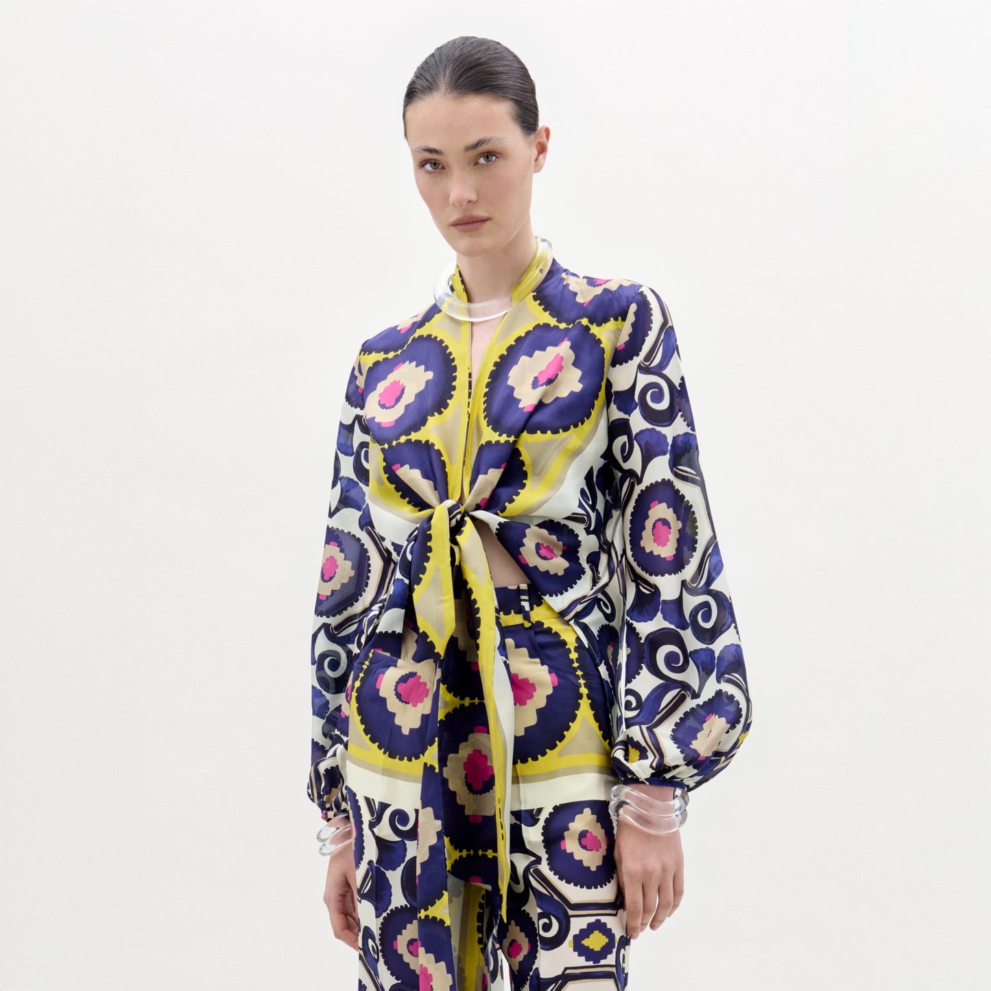 Una persona aparece sobre un fondo blanco liso con la blusa Honey, un top de manga larga de seda 100% con estampados circulares y florales en morado, amarillo, azul marino y rosa. Tiene mangas abullonadas y un lazo delantero. Fabricada en Colombia.