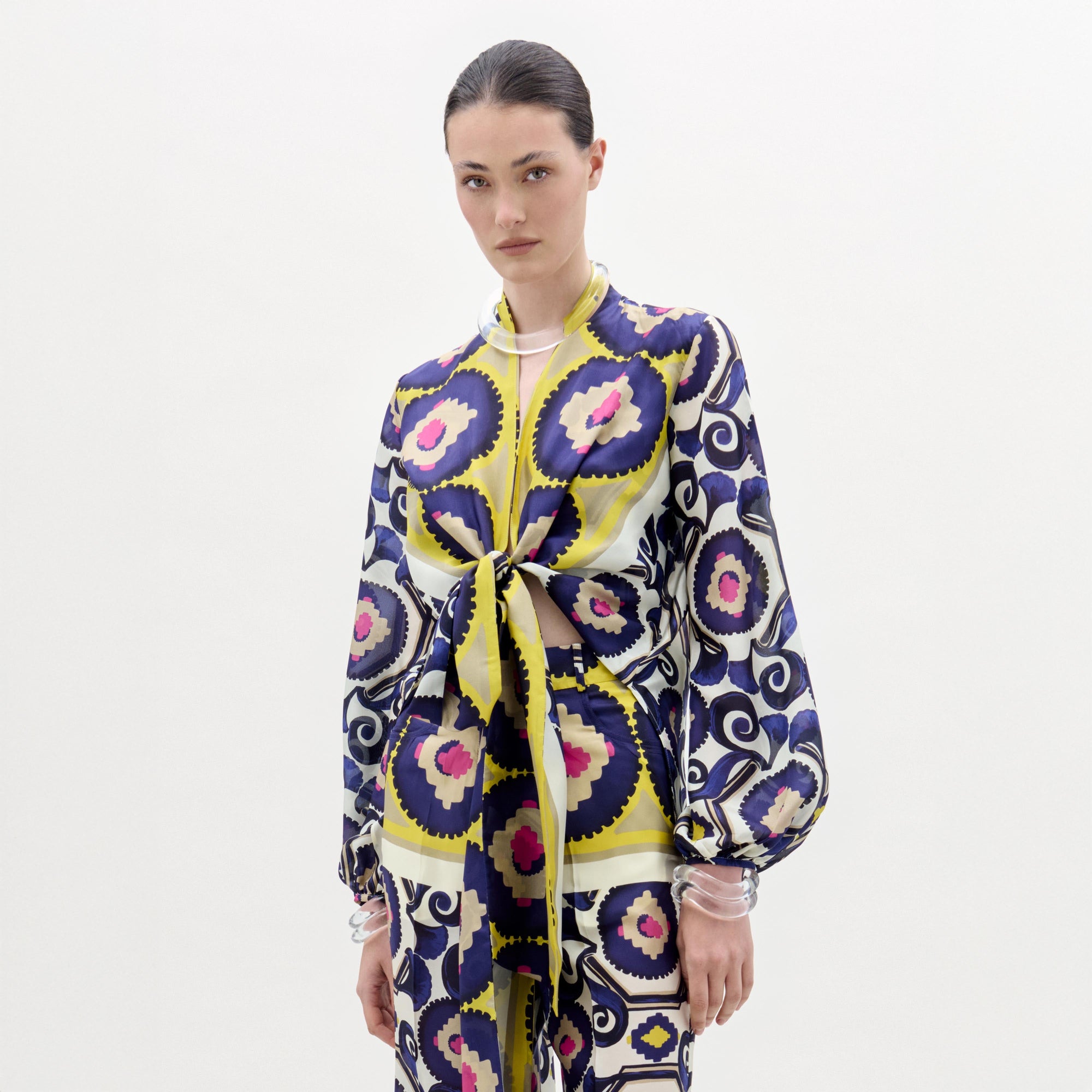 Una persona aparece sobre un fondo blanco liso con la blusa Honey, un top de manga larga de seda 100% con estampados circulares y florales en morado, amarillo, azul marino y rosa. Tiene mangas abullonadas y un lazo delantero. Fabricada en Colombia.