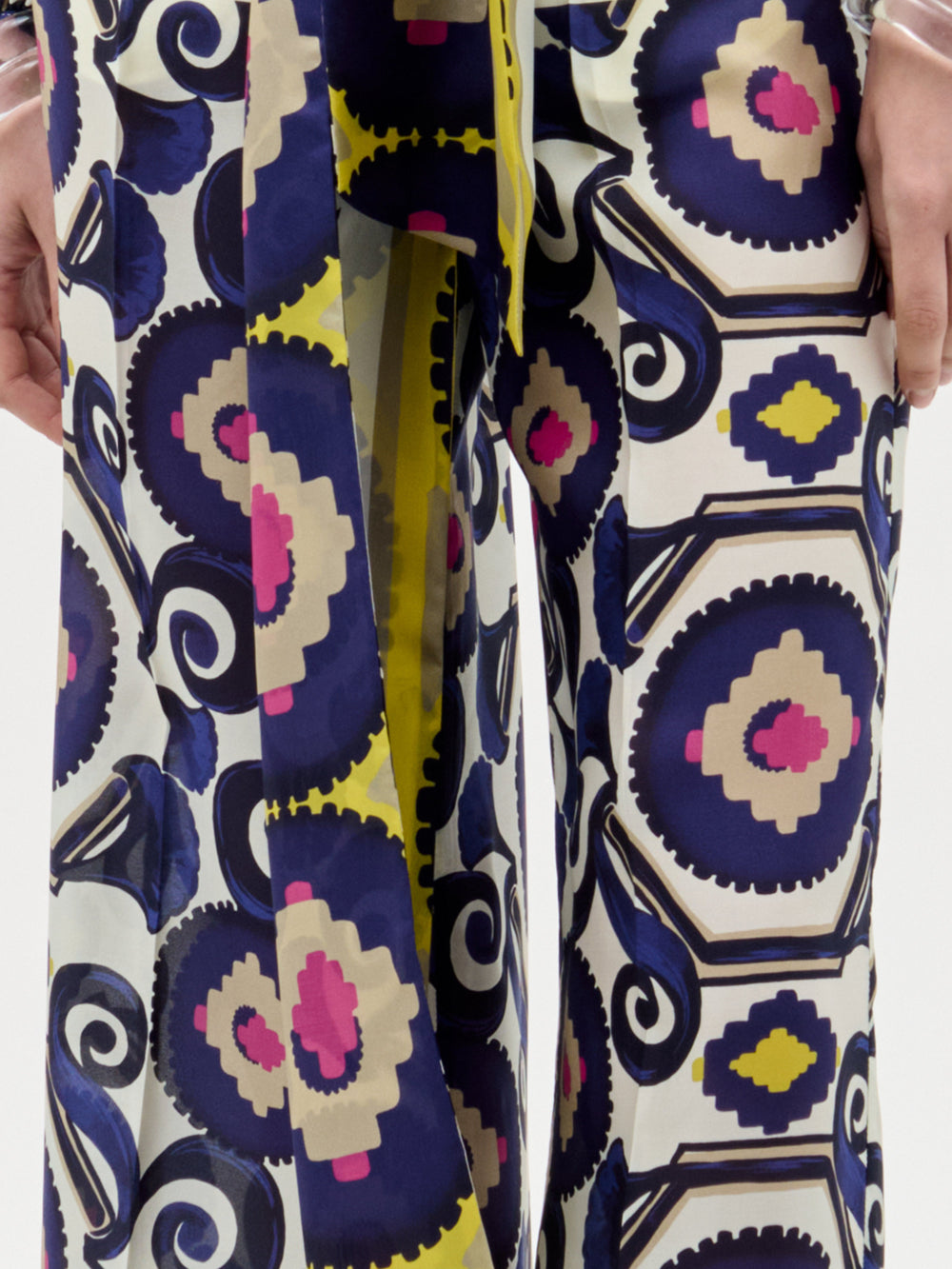 Primer plano de las piernas de una persona que lleva el pantalón Emine Pant, con un atrevido estampado geométrico en azul marino, crema, rosa y amarillo. El corte bota ancha y las manos parcialmente visibles aportan un estilo moderno a este llamativo look.