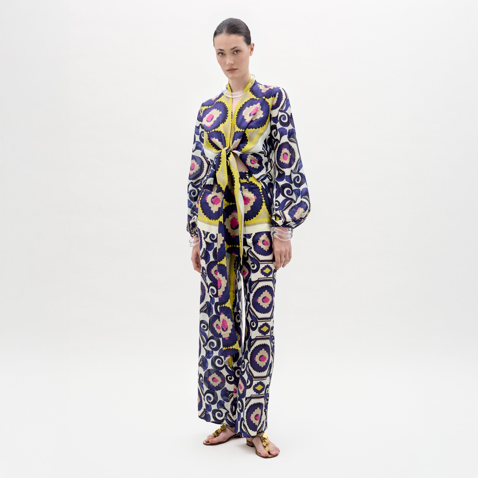 Sobre un fondo blanco liso, una persona lleva el pantalón Emine, largo y entero, con un llamativo dibujo geométrico en azul, amarillo, rosa y blanco. Lleva el pelo recogido y calza sandalias.