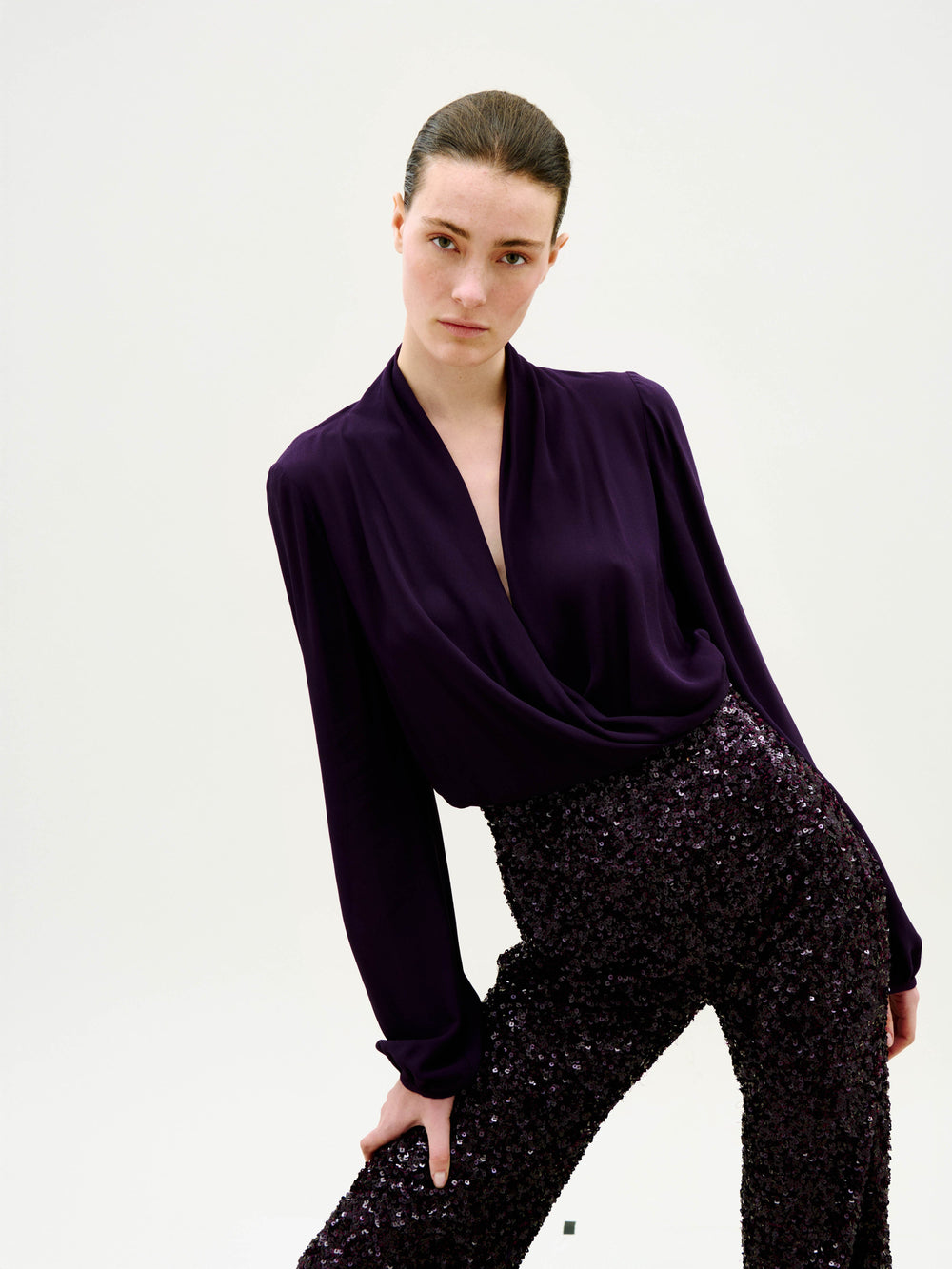 A person poses against a white background, wearing high-waisted Avellino Pants with dark sequins and a deep purple, long-sleeved blouse featuring a draped neckline.