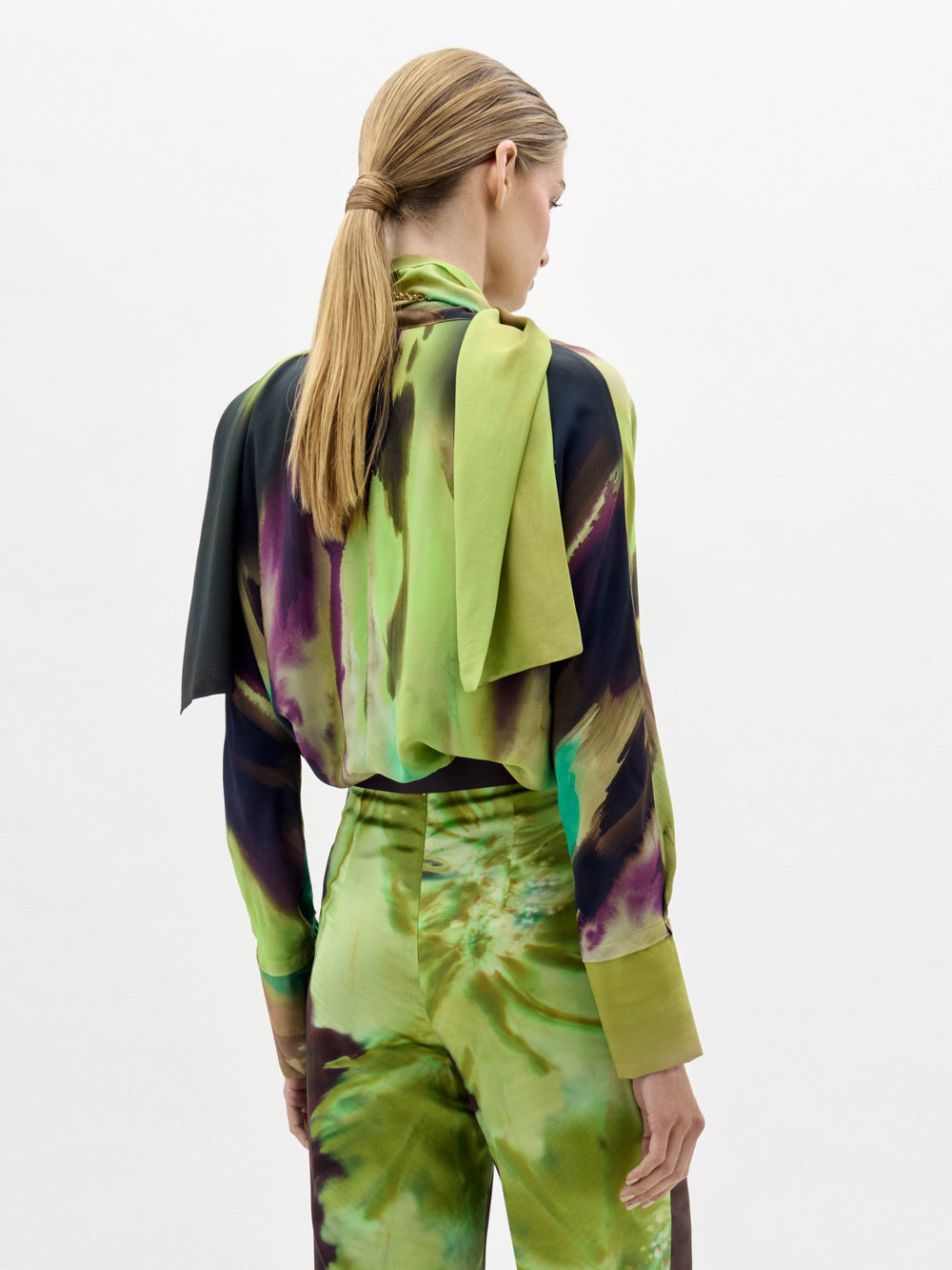 Una persona con el pelo rubio, largo y liso, recogido en una coleta baja, aparece de espaldas con la blusa Jaboca, un conjunto estampado en verde, morado y negro con un detalle drapeado sobre un hombro. El fondo es blanco.