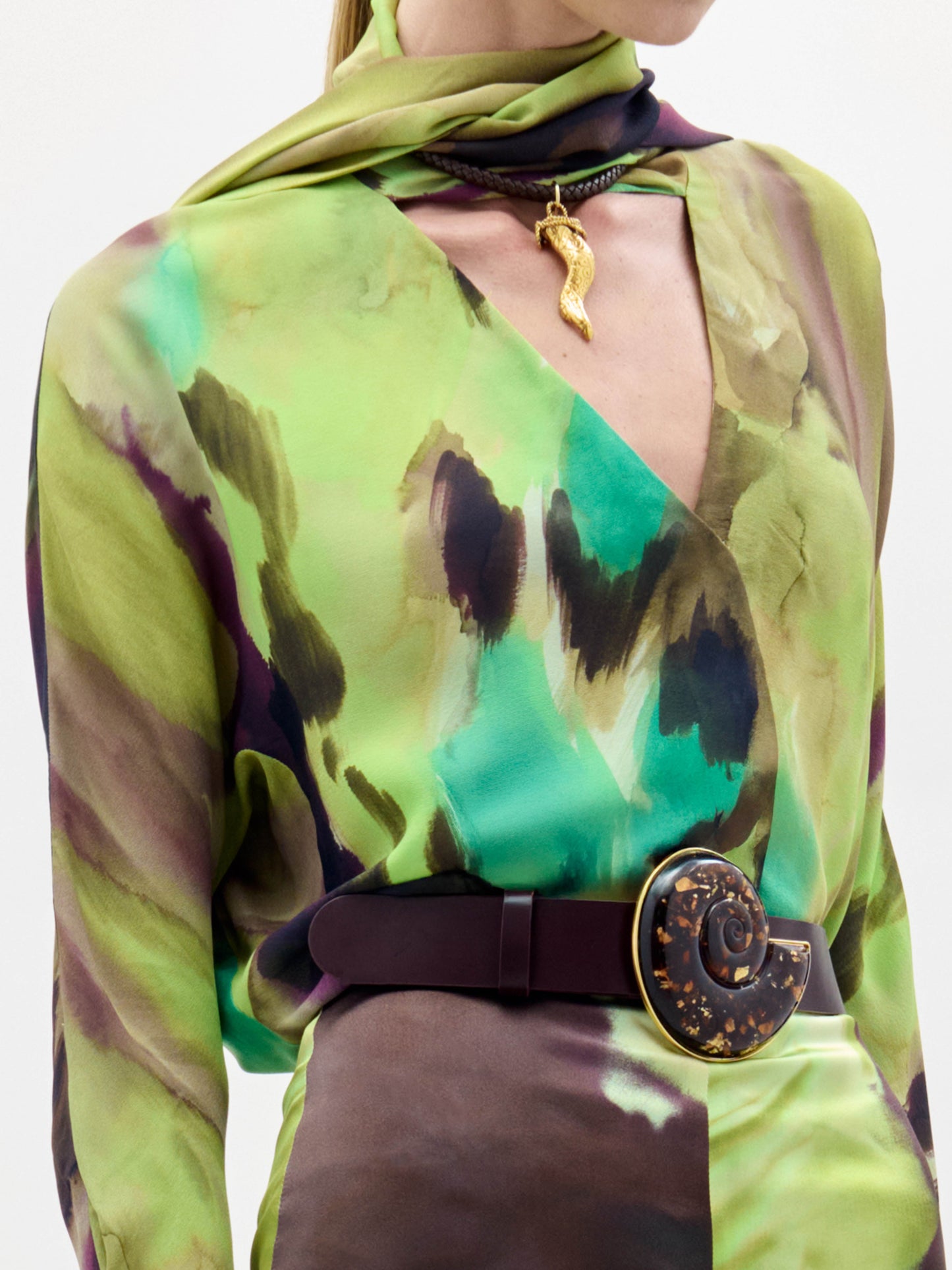 Una persona lleva la blusa Jaboca con motivos abstractos verdes y marrones, combinada con un pañuelo a juego, un collar con colgante dorado y un cinturón ancho oscuro con una gran hebilla circular texturizada.