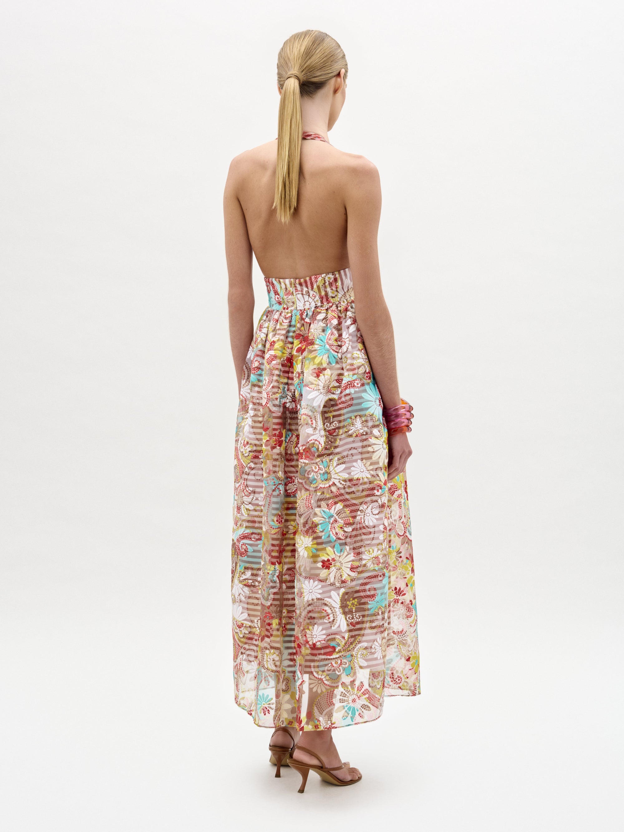 Una mujer rubia con coleta mira hacia otro lado y lleva el vestido Kairi, sin tirantes y hasta los tobillos, con un colorido estampado floral, y sandalias de tacón alto. Lleva un bolso en la mano derecha y brazaletes rosas en la muñeca izquierda.