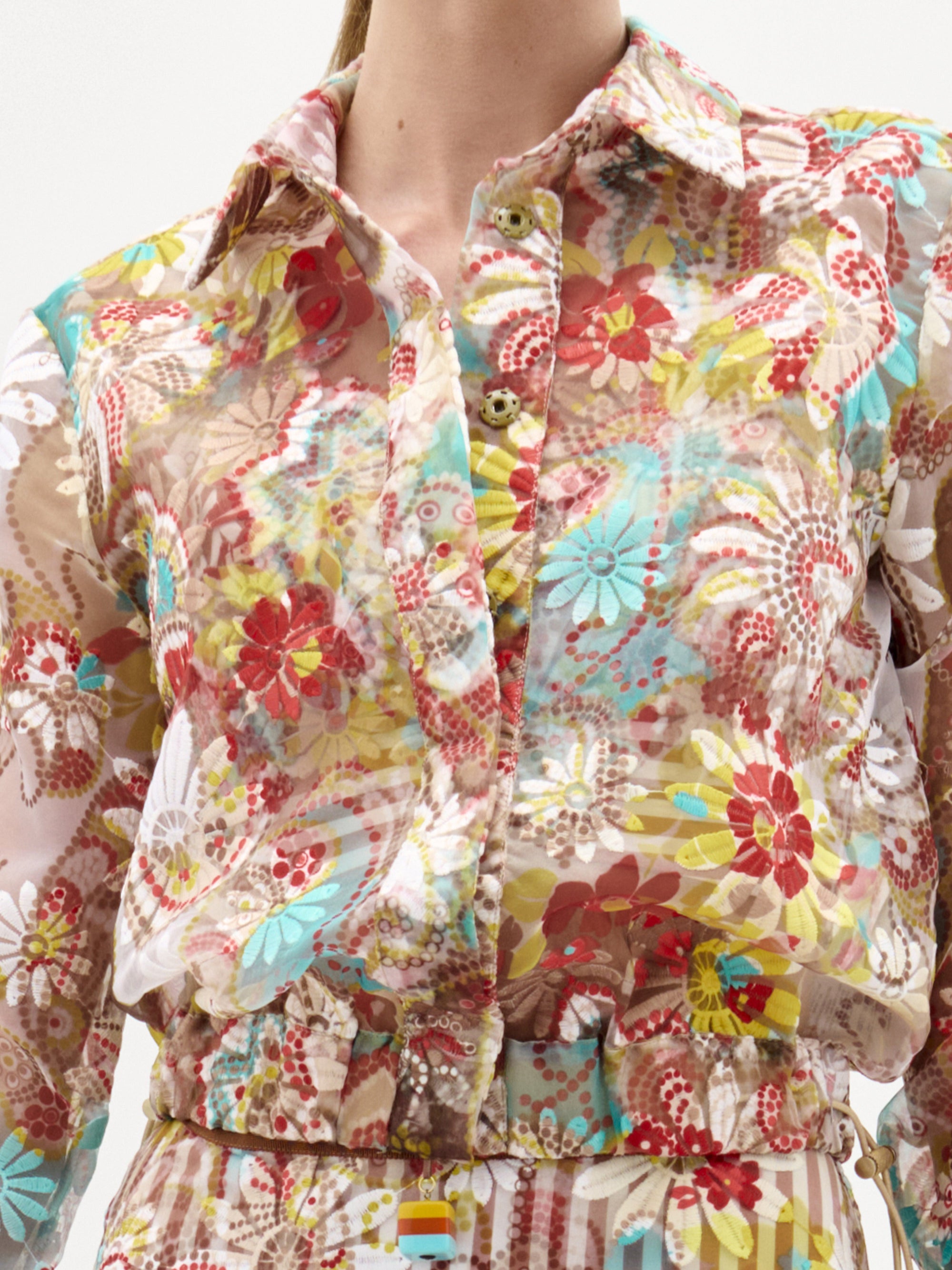 Una persona lleva la Chaqueta Katniss, una blusa transparente de manga larga confeccionada en tela italiana con un estampado floral y geométrico multicolor en rojo, amarillo, azul y beige. Tiene cuello y botones delanteros.