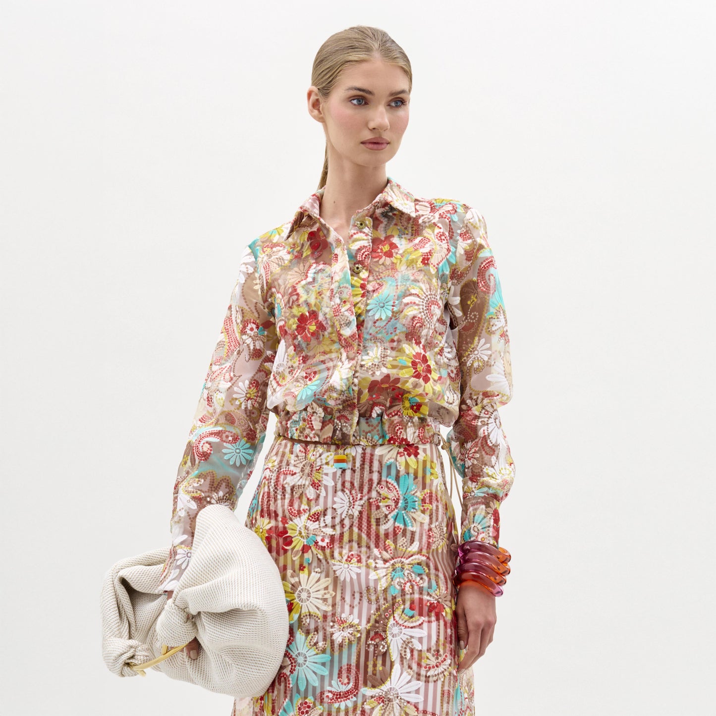 Una mujer, sobre un fondo liso, lleva la chaqueta Katniss -una blusa de manga larga y estampado floral- y una falda a juego. Lleva un gran bolso blanco con textura y brazaletes translúcidos apilados en la muñeca derecha.
