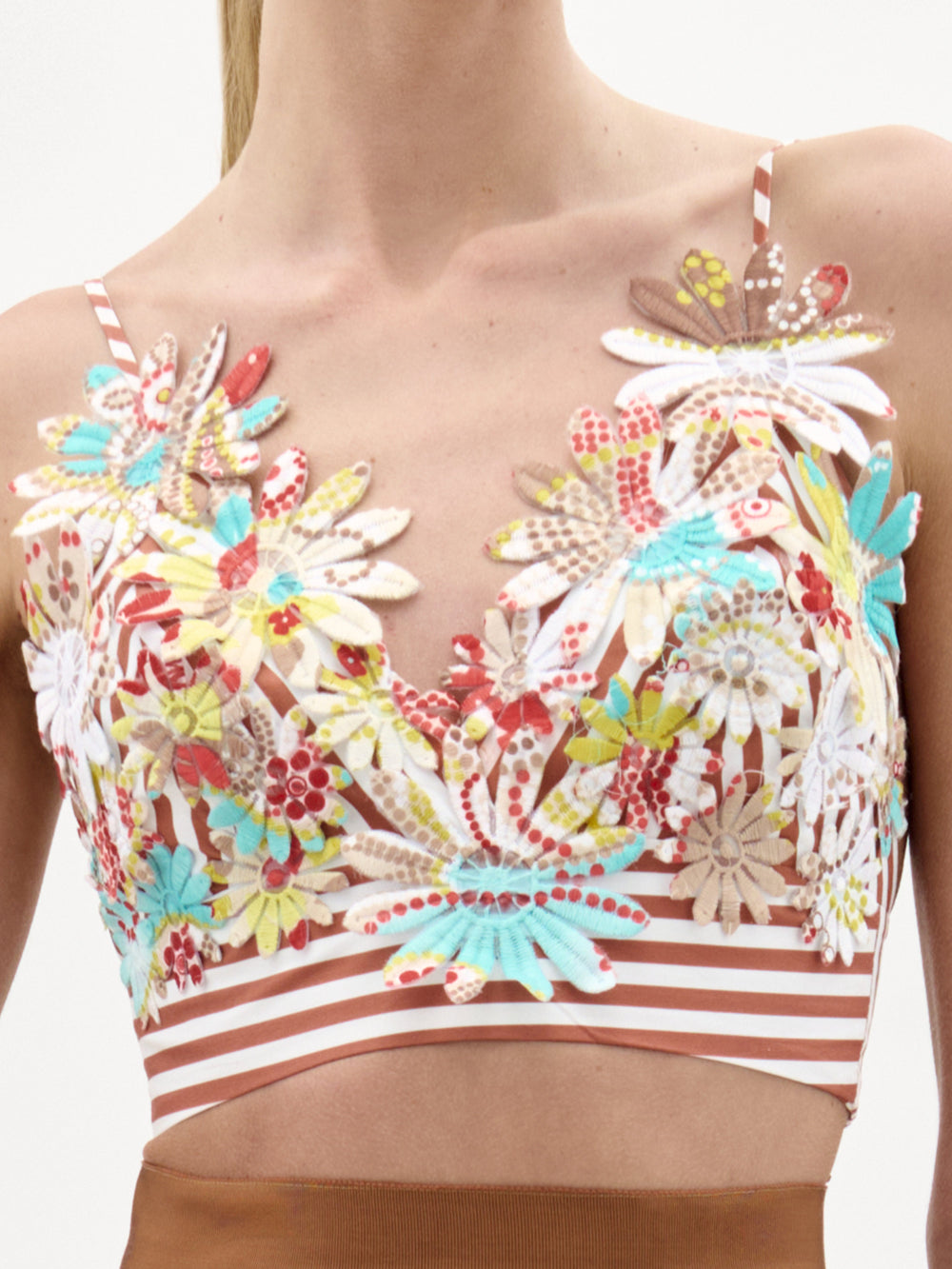 Primer plano de una mujer que lleva el Ibbie Top, un colorido top con cuello en V y apliques florales, tirantes finos y una banda a rayas marrones y blancas bajo el pecho, fabricado en Colombia. El fondo es liso y de color claro.