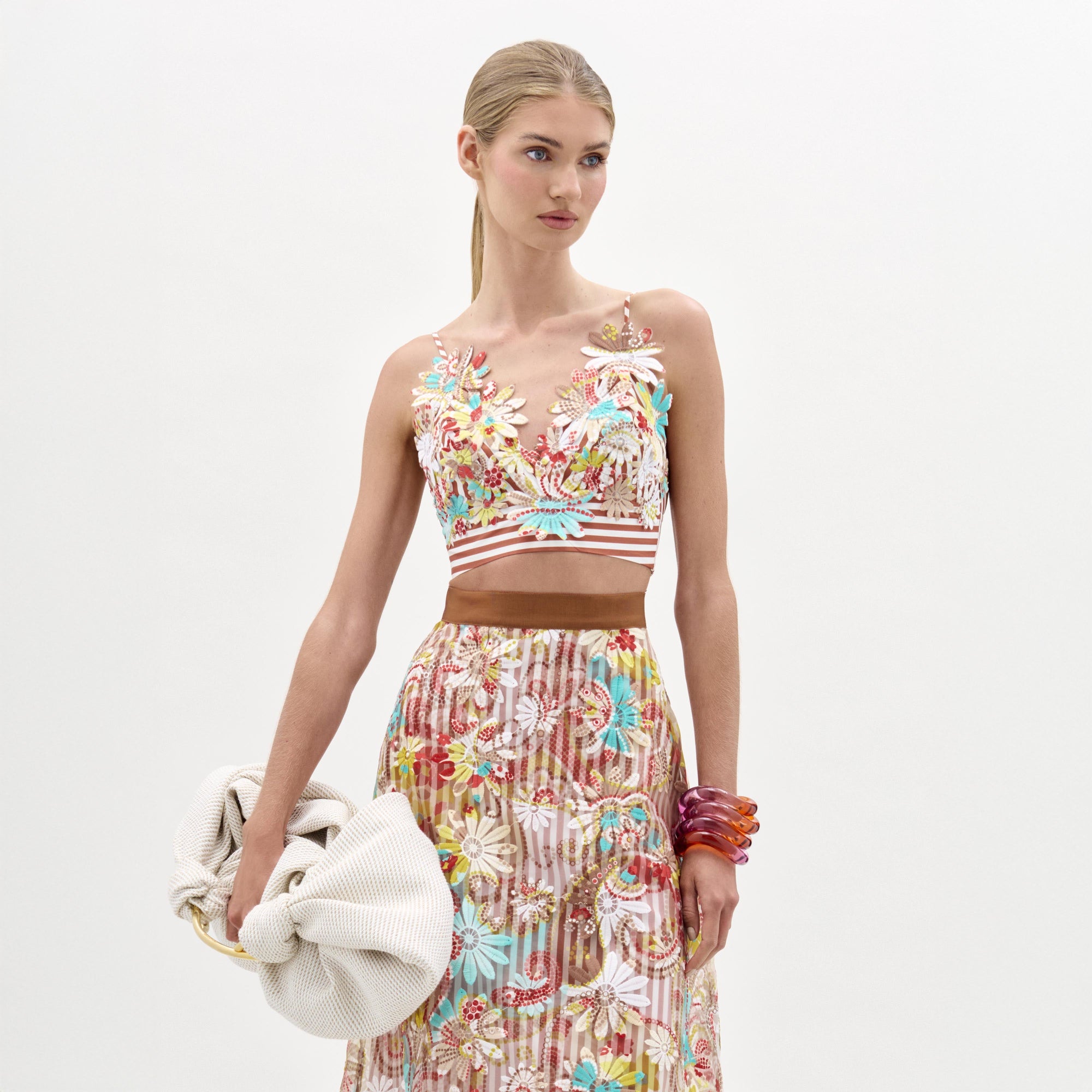 Una mujer sobre un fondo liso luce el top Ibbie, un top bordado de flores con cuello en V y falda a juego. Lleva un bolso blanco grande con textura, pulseras rojas gruesas en la muñeca izquierda y este look está hecho en Colombia.