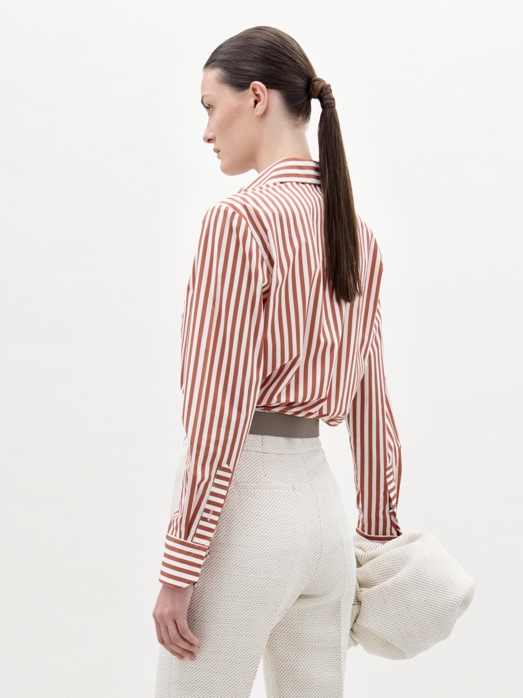 Una mujer con el pelo castaño liso recogido en una coleta baja aparece de perfil y de espaldas, con la blusa Karole -una blusa abotonada a rayas rojas y blancas- metida por dentro de unos pantalones blancos de cintura alta, sosteniendo una prenda blanca texturizada en la mano izquierda.