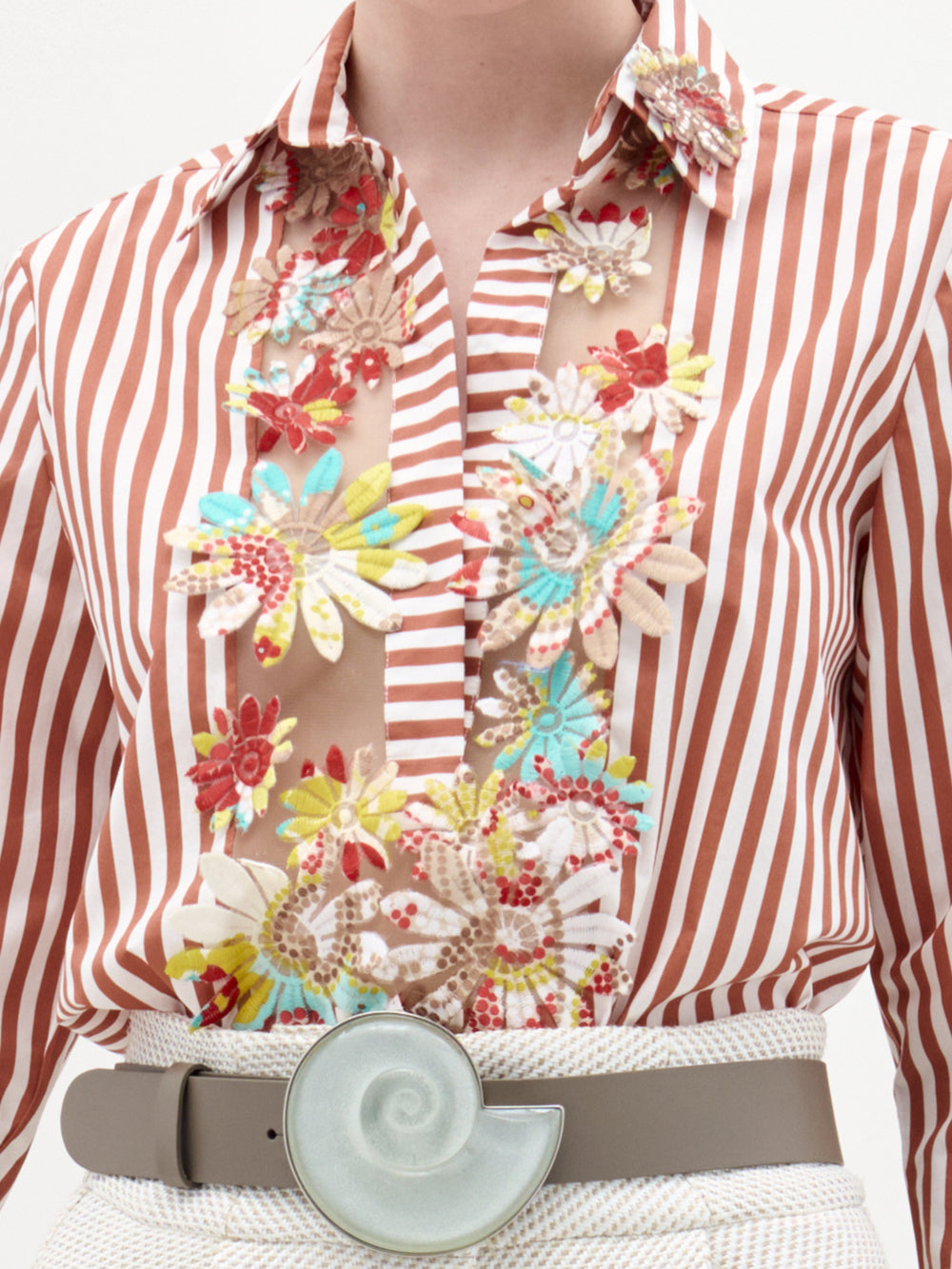 Una persona lleva la blusa Karole, con rayas verticales rojas y blancas, paneles transparentes y aplicaciones florales bordadas, combinada con un cinturón beige con una gran hebilla redonda en forma de concha de caracol.