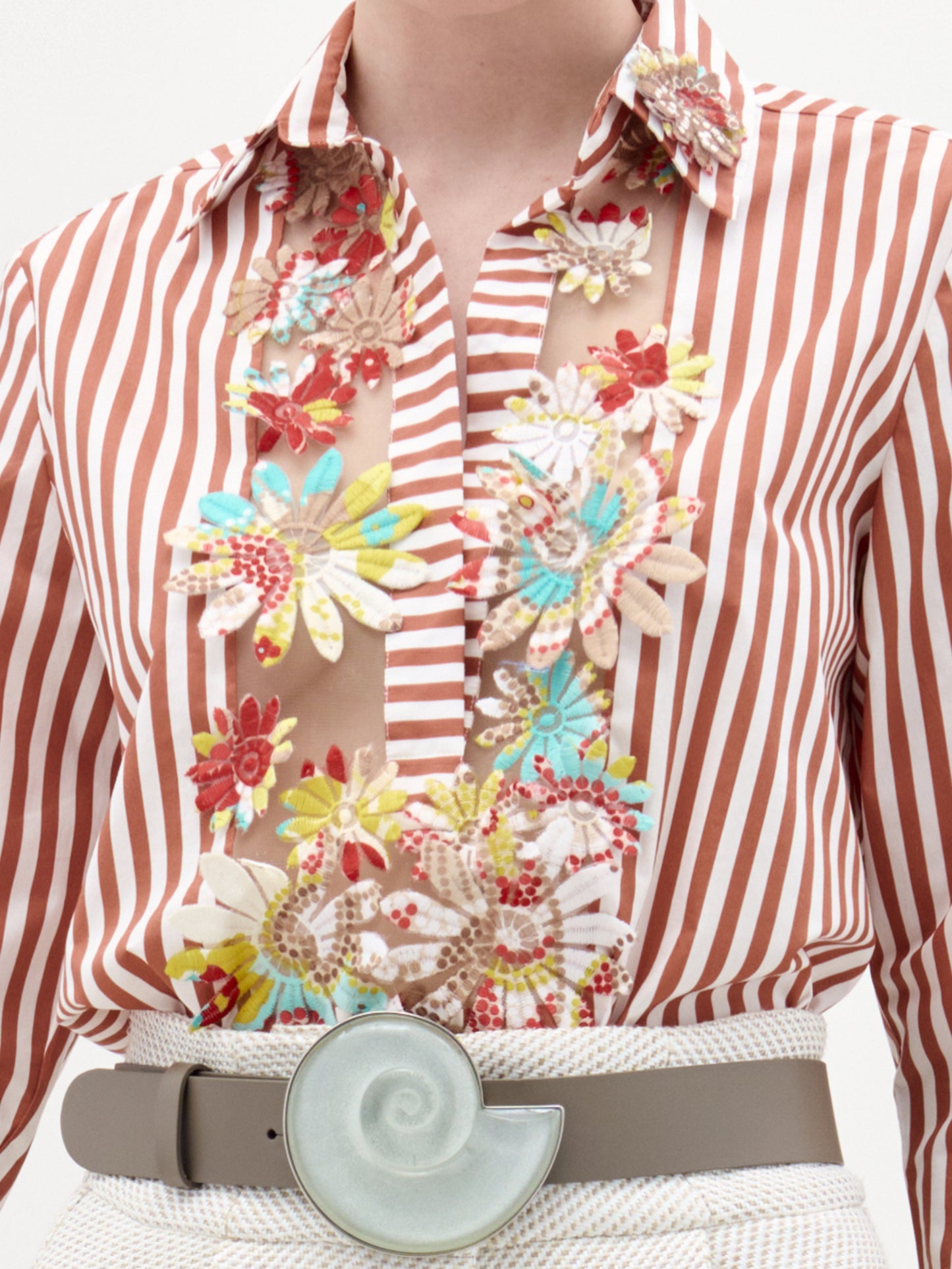 Una persona lleva la blusa Karole, con rayas verticales rojas y blancas, paneles transparentes y aplicaciones florales bordadas, combinada con un cinturón beige con una gran hebilla redonda en forma de concha de caracol.