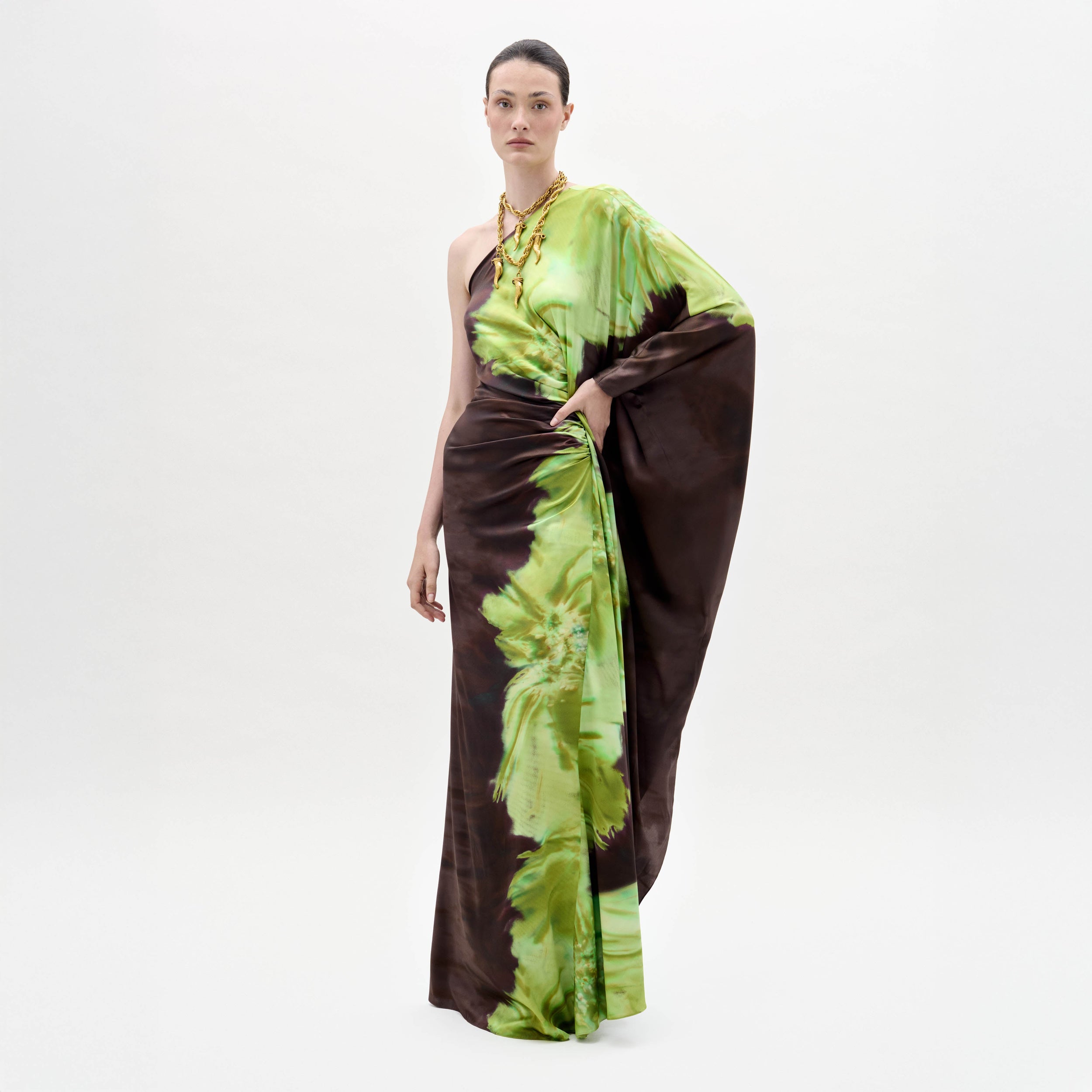 Una mujer sobre un fondo blanco liso luce el vestido Afrodita, de un solo hombro y largo hasta el suelo, con un estampado abstracto en tonos marrones y verdes. Lleva el pelo recogido y accesorios como collares dorados.