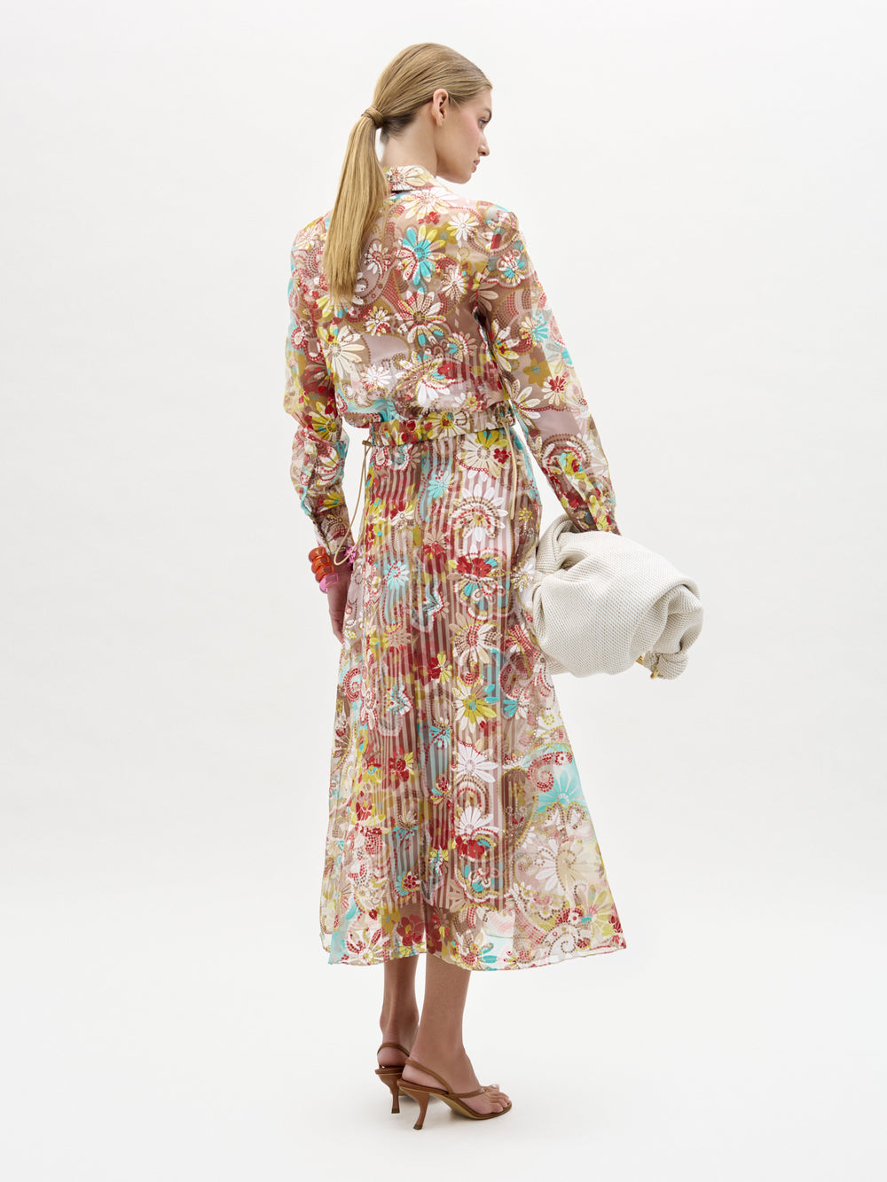 Una mujer de espaldas lleva la falda Nelida, una falda midi larga de flores transparentes y manga larga, y un bolso de mano grande con textura de color hueso. Lleva el pelo castaño claro recogido en una coleta baja y combina el conjunto con sandalias de tacón marrones.