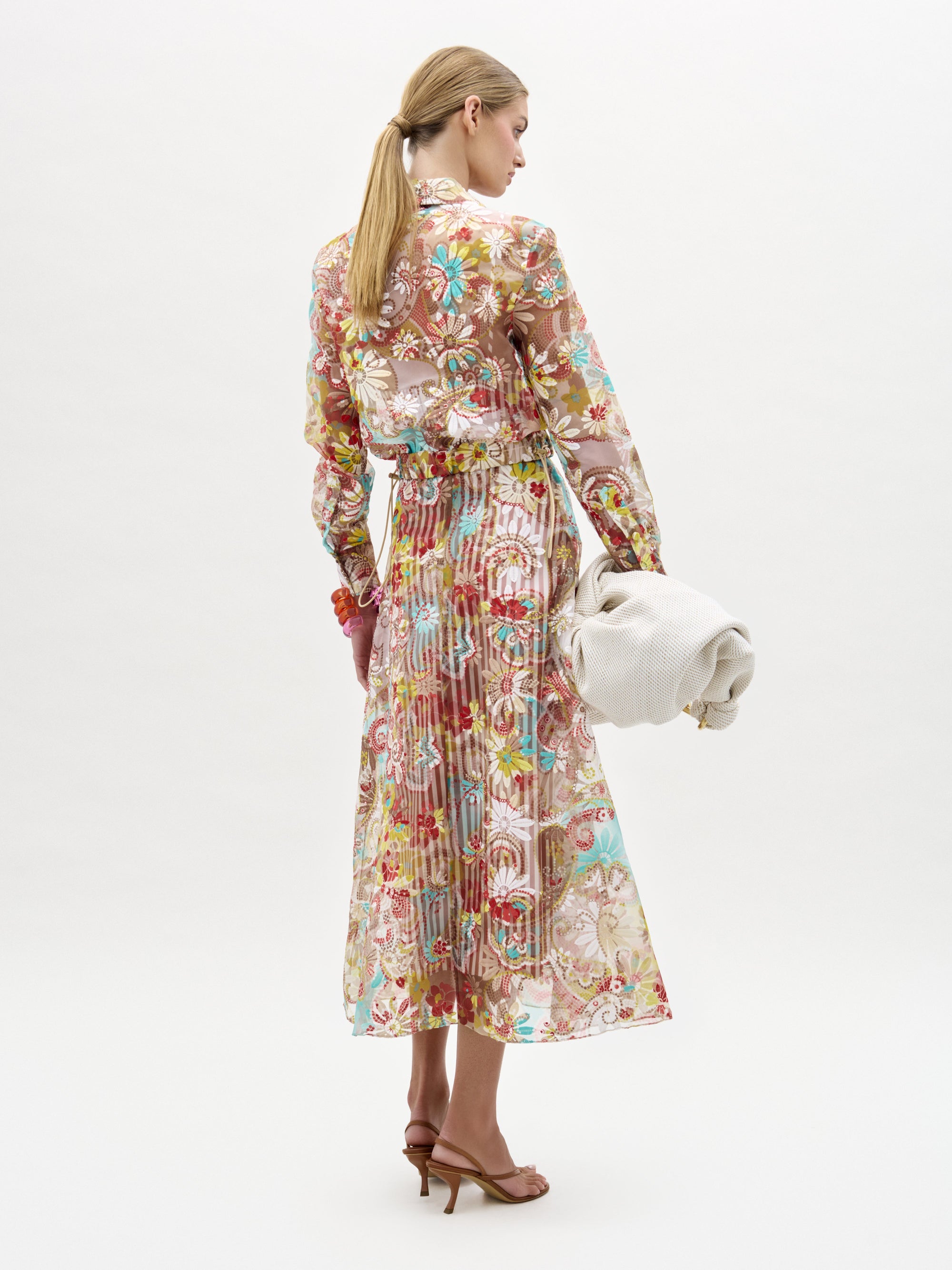 Una mujer de espaldas lleva la falda Nelida, una falda midi larga de flores transparentes y manga larga, y un bolso de mano grande con textura de color hueso. Lleva el pelo castaño claro recogido en una coleta baja y combina el conjunto con sandalias de tacón marrones.
