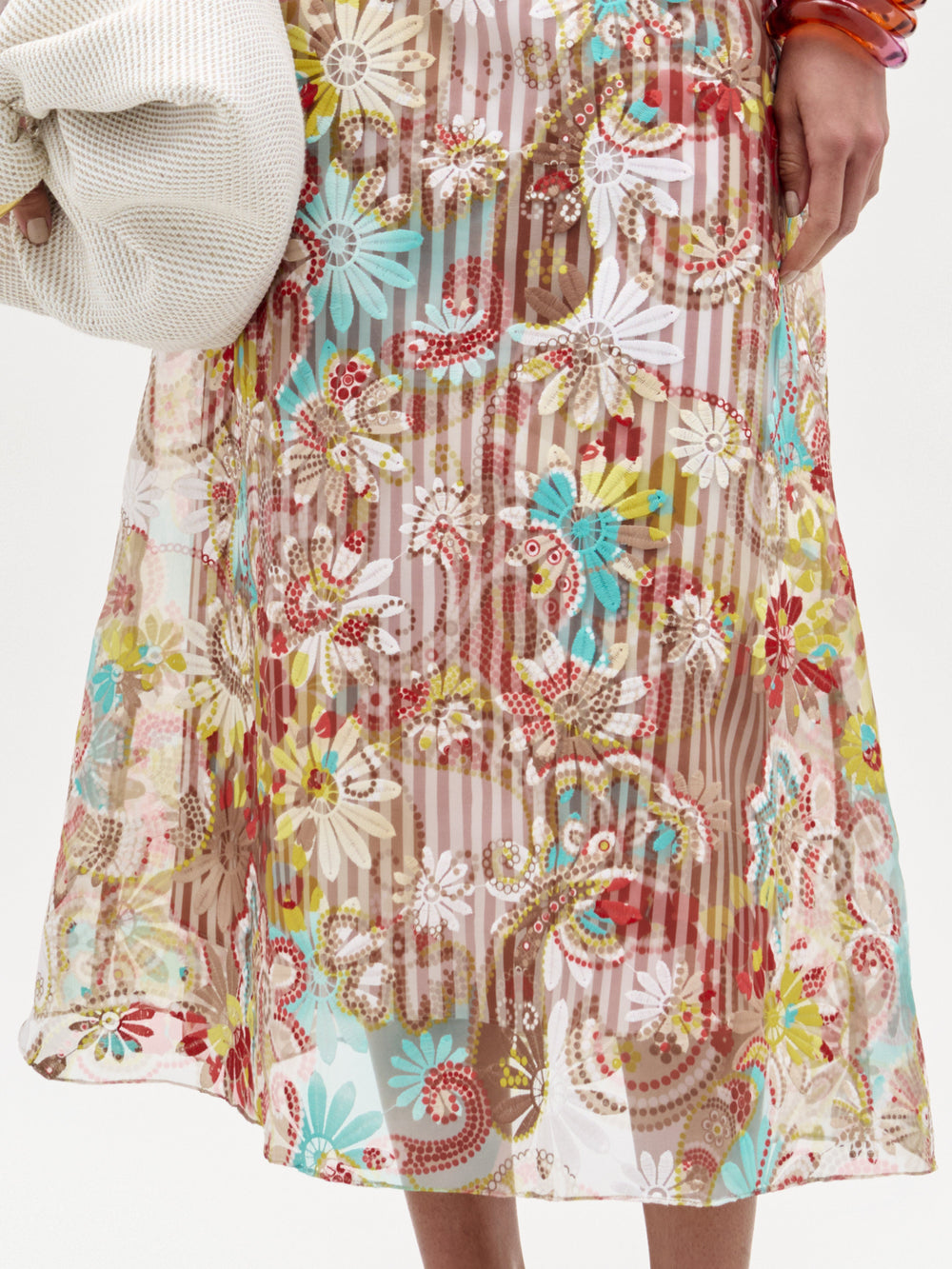 Una persona lleva la falda Nelida -con coloridos motivos florales y paisley sobre rayas verticales-, confeccionada en Colombia. Solo se ven la parte inferior del torso y la mano que sujeta un tejido blanco texturizado.