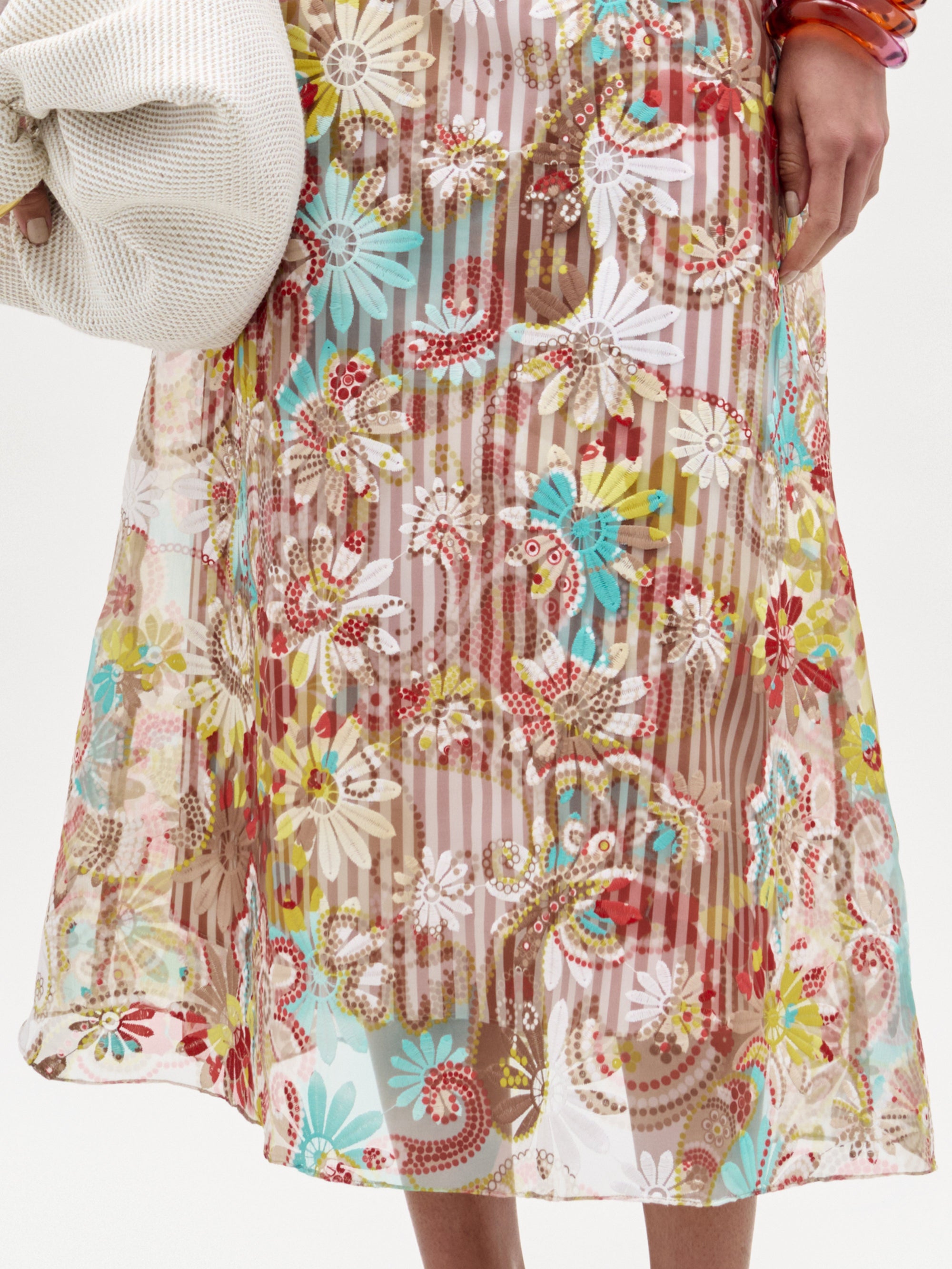 Una persona lleva la falda Nelida -con coloridos motivos florales y paisley sobre rayas verticales-, confeccionada en Colombia. Solo se ven la parte inferior del torso y la mano que sujeta un tejido blanco texturizado.