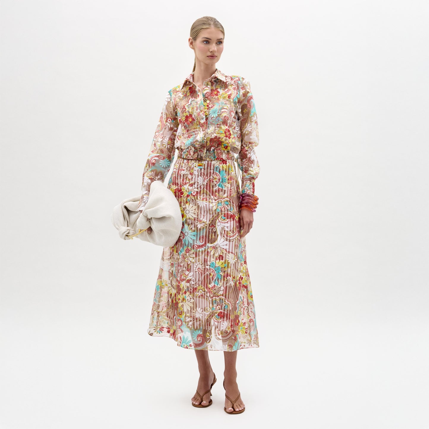 Sobre un fondo blanco, una mujer lleva una blusa de flores de manga larga y la falda Nelida, ambas confeccionadas en Colombia, combinadas con sandalias de tacón marrones, brazaletes de colores y un bolso de mano grande de color claro.