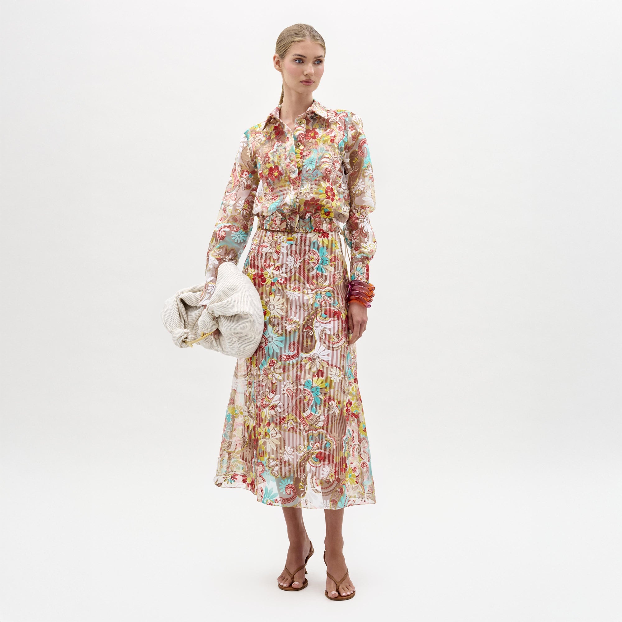 Sobre un fondo blanco, una mujer lleva una blusa de flores de manga larga y la falda Nelida, ambas confeccionadas en Colombia, combinadas con sandalias de tacón marrones, brazaletes de colores y un bolso de mano grande de color claro.