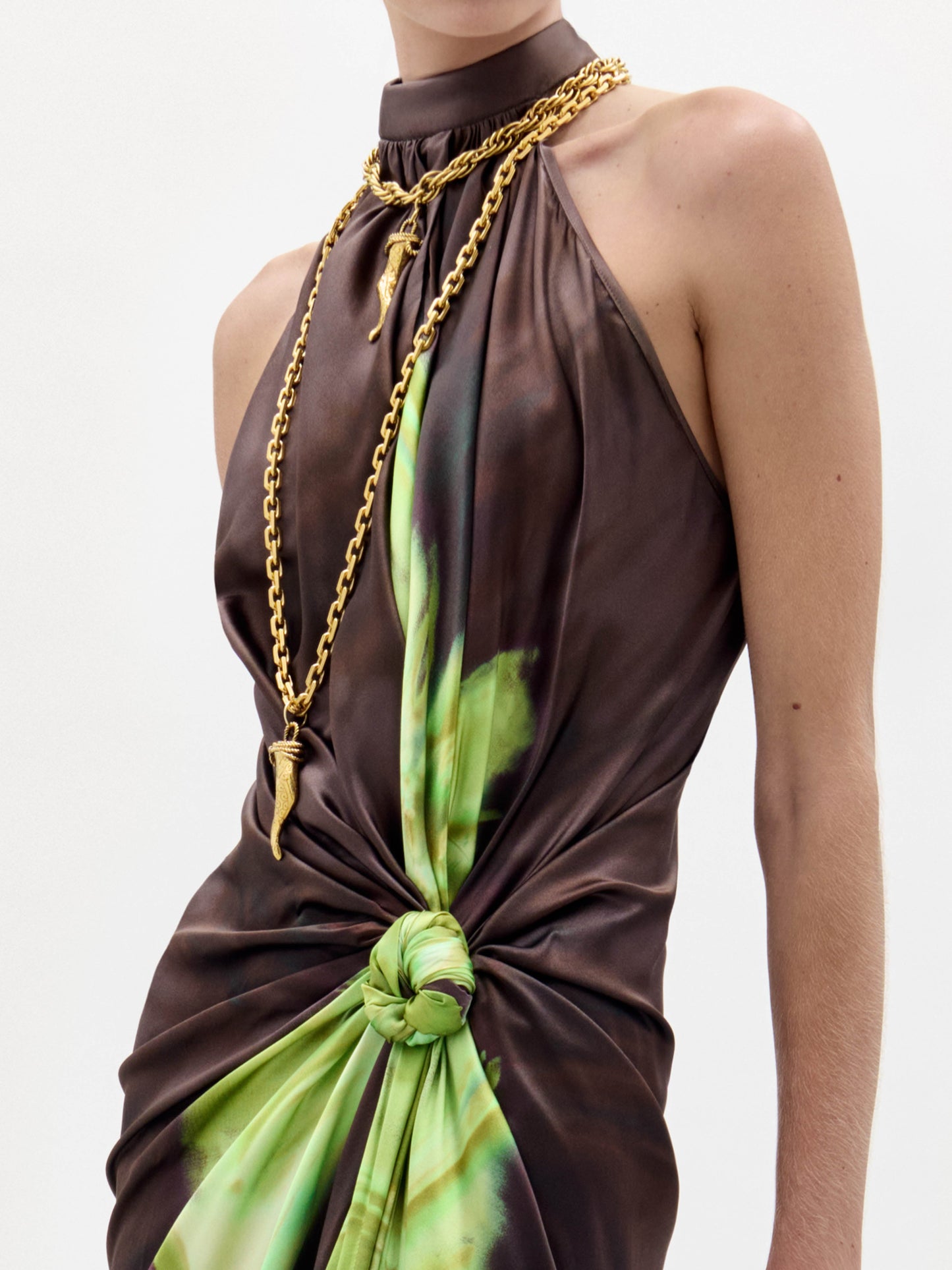 Una persona lleva el Vestido Tizziana -un vestido anudado sin mangas, marrón y verde, con escote alto- combinado con dos largos collares de cadena de oro con colgantes sobre un fondo liso y claro.