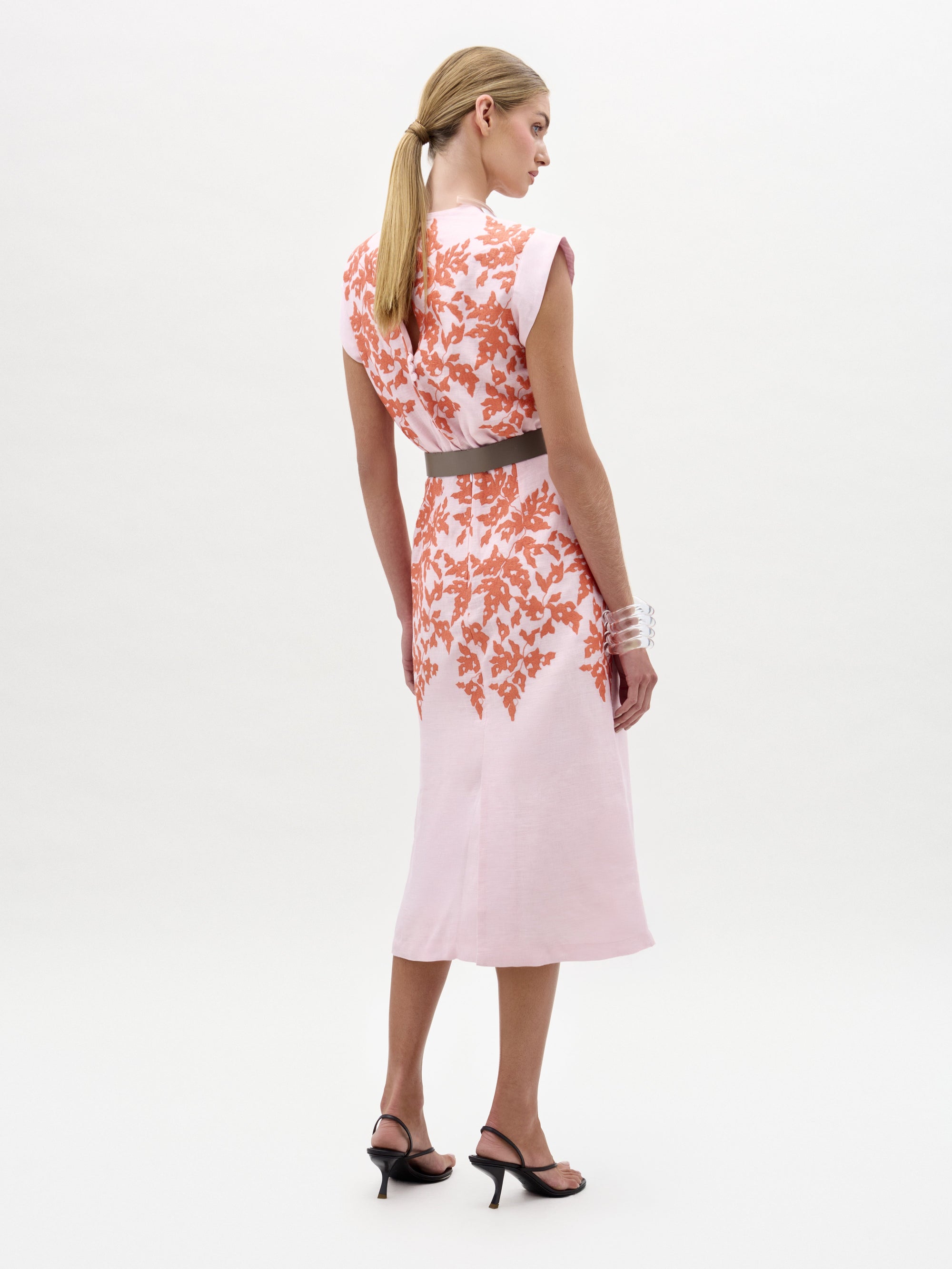 Una mujer con el pelo rubio recogido en una coleta baja mira hacia otro lado y lleva el Vestido Oliva -un vestido midi rosa claro con motivos florales naranjas- combinado con sandalias de tacón negras, un cinturón gris y una pulsera blanca.