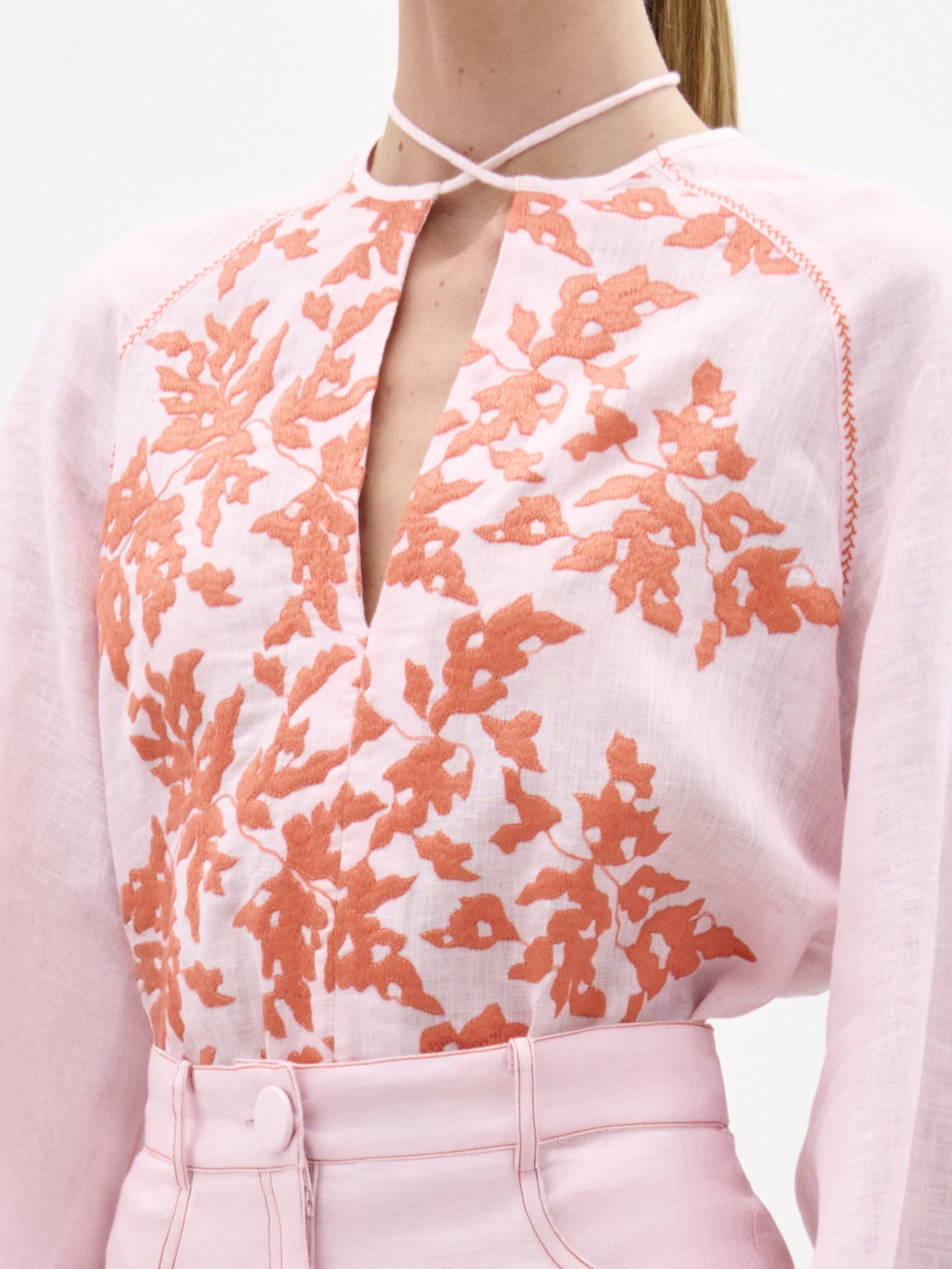 Una persona lleva la blusa Molveno, un top de lino rosa claro con bordados florales naranjas y escote en pico. Solo se ve la parte superior del cuerpo, combinada con un pantalón rosa claro de cintura alta.