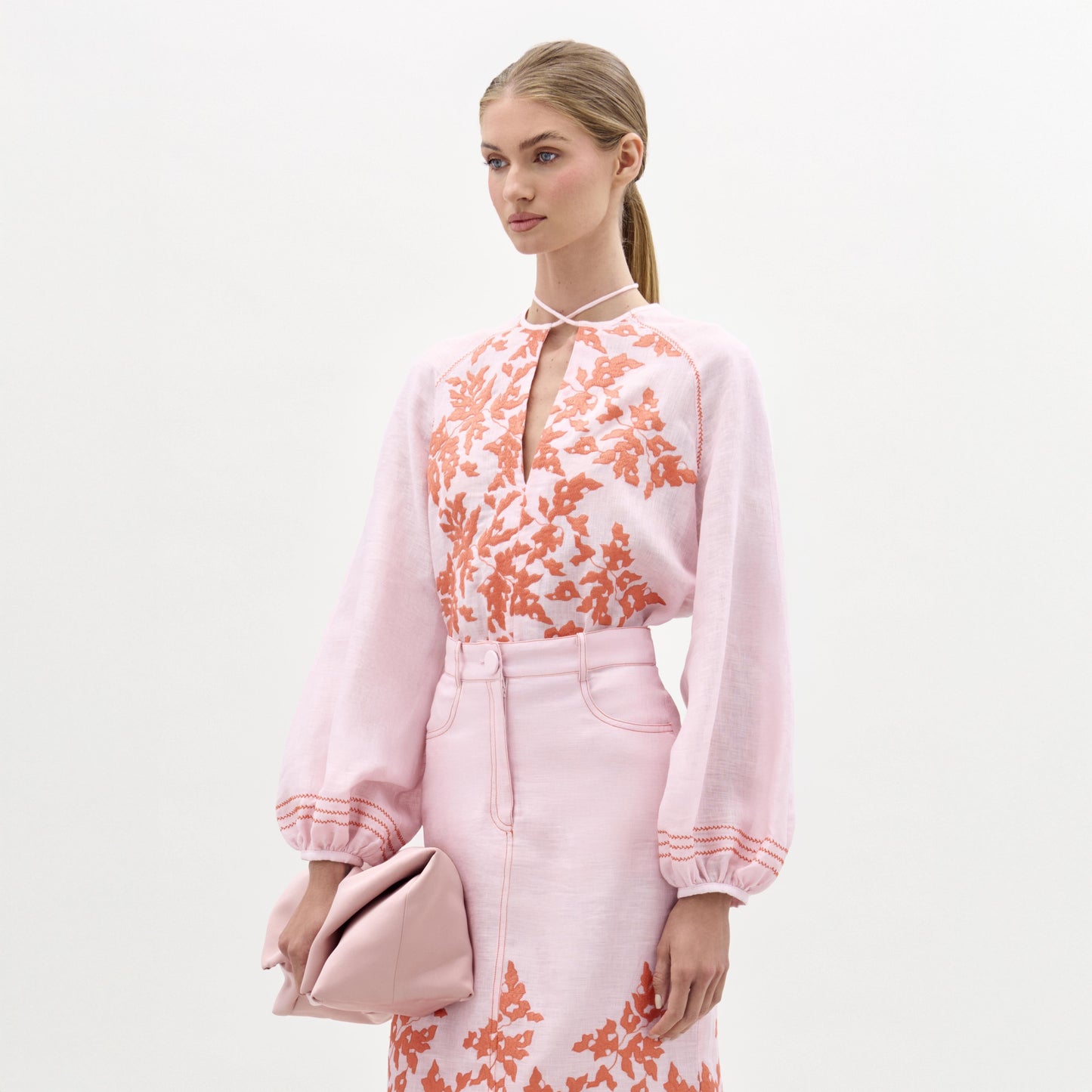 Una mujer lleva la blusa Molveno -de lino rosa claro con bordados florales naranjas y escote en V- de pie sobre un fondo blanco liso, sujeta un bolso de mano rosa a juego y mira ligeramente hacia un lado.